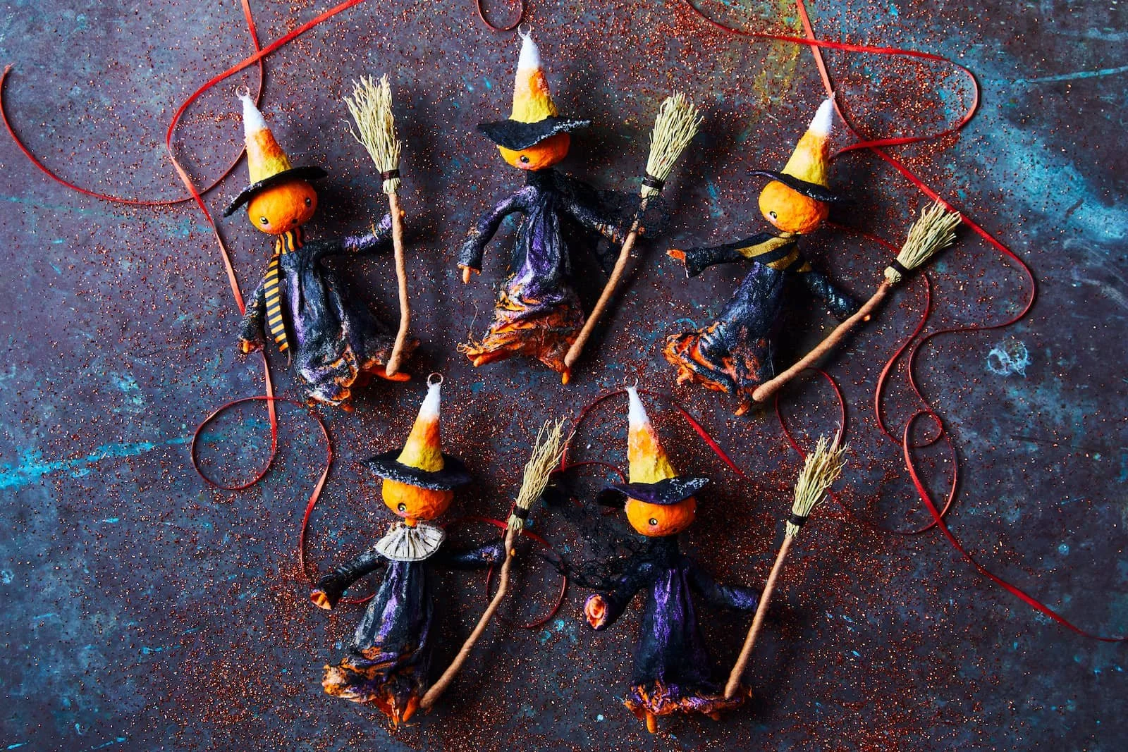 Set of handmade Halloween witch decorations with pumpkin heads, witch hats, black cloaks, broomsticks, and red strings, arranged on a colorful textured surface.