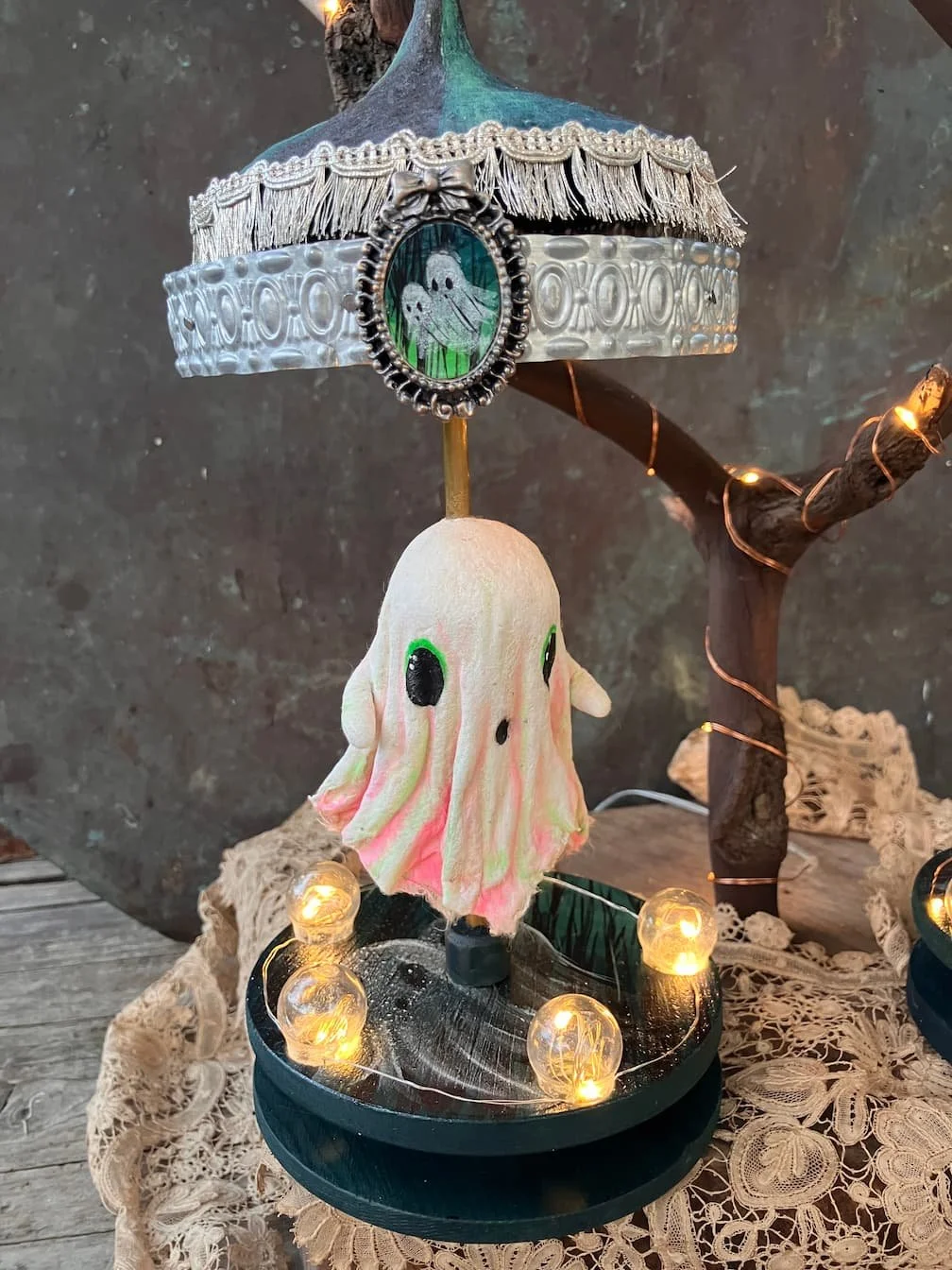 A Halloween sculpted lamp featuring a small ghost figure with green eyes, a decorative frame with a spooky face, a fringed lampshade, and string lights, all arranged on a wooden surface with lace fabric and a twisted branches as background.