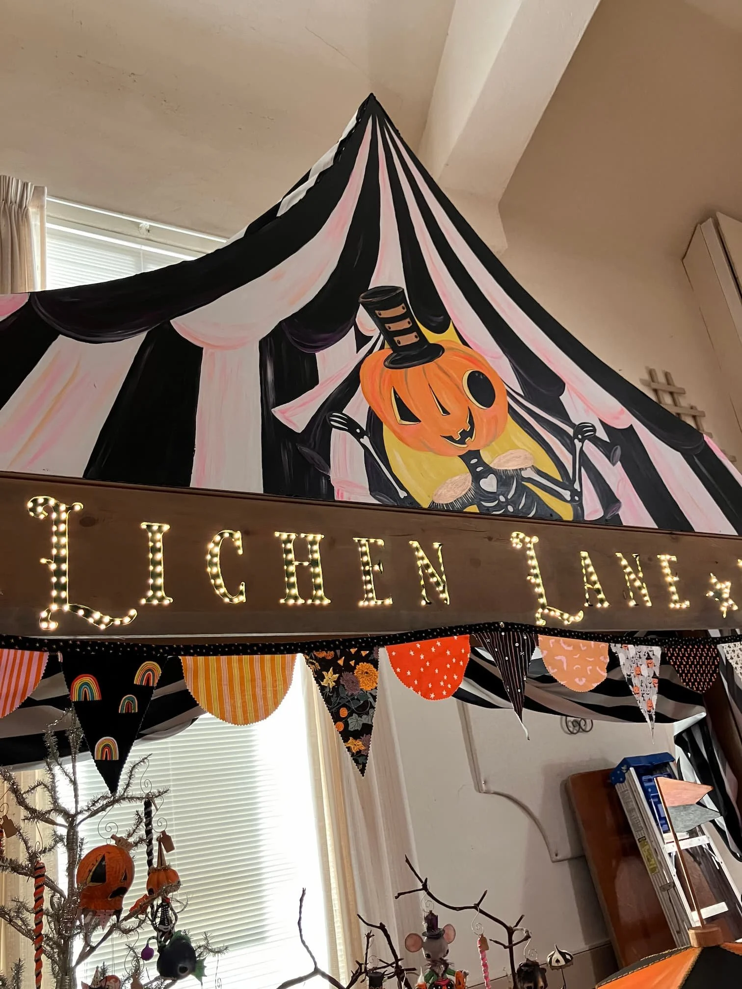 Decorated room with Halloween and fall theme decorations, including a banner with various colors and patterns, string lights forming the word 'LUCKEN,' a pumpkin face illustration on the wall, and holiday-themed ornaments hanging from a small tree.