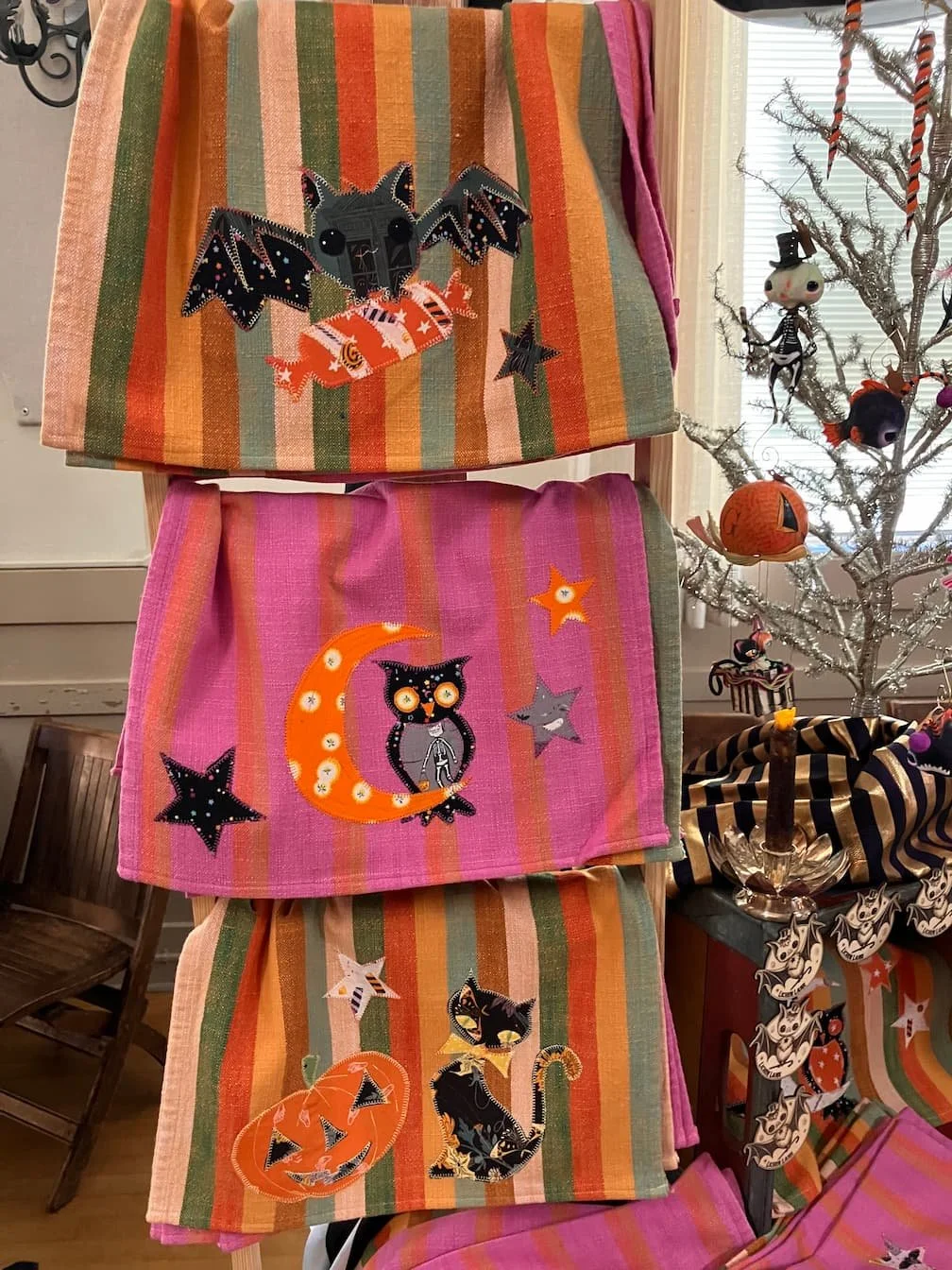 Decorative Halloween tea towels with embroidered black cats, pumpkins, and stars displayed on a chair and table with Halloween themed ornaments, including a skeleton, a pumpkin, and bats.