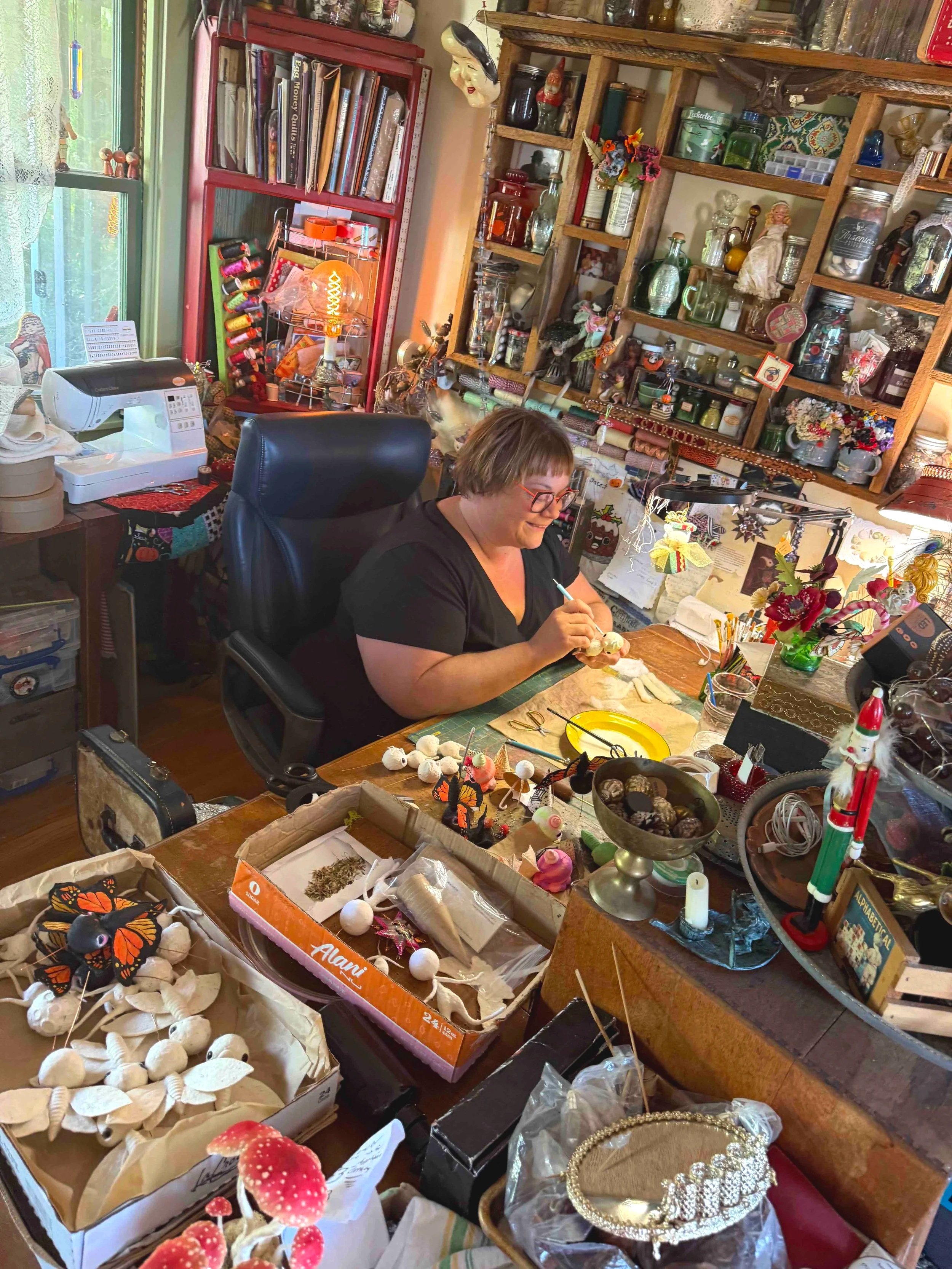 A cluttered craft or flea market table filled with various decorative items, jewelry, small figurines, artificial mushrooms, and craft supplies, with a person seated working on a project in a home or shop setting.