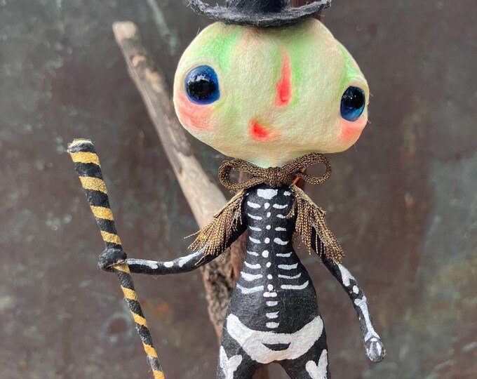 A whimsical handmade doll with a painted face, wearing a black and white skeleton costume, and a bow. The doll is holding a striped stick and has a tree branch in the background.