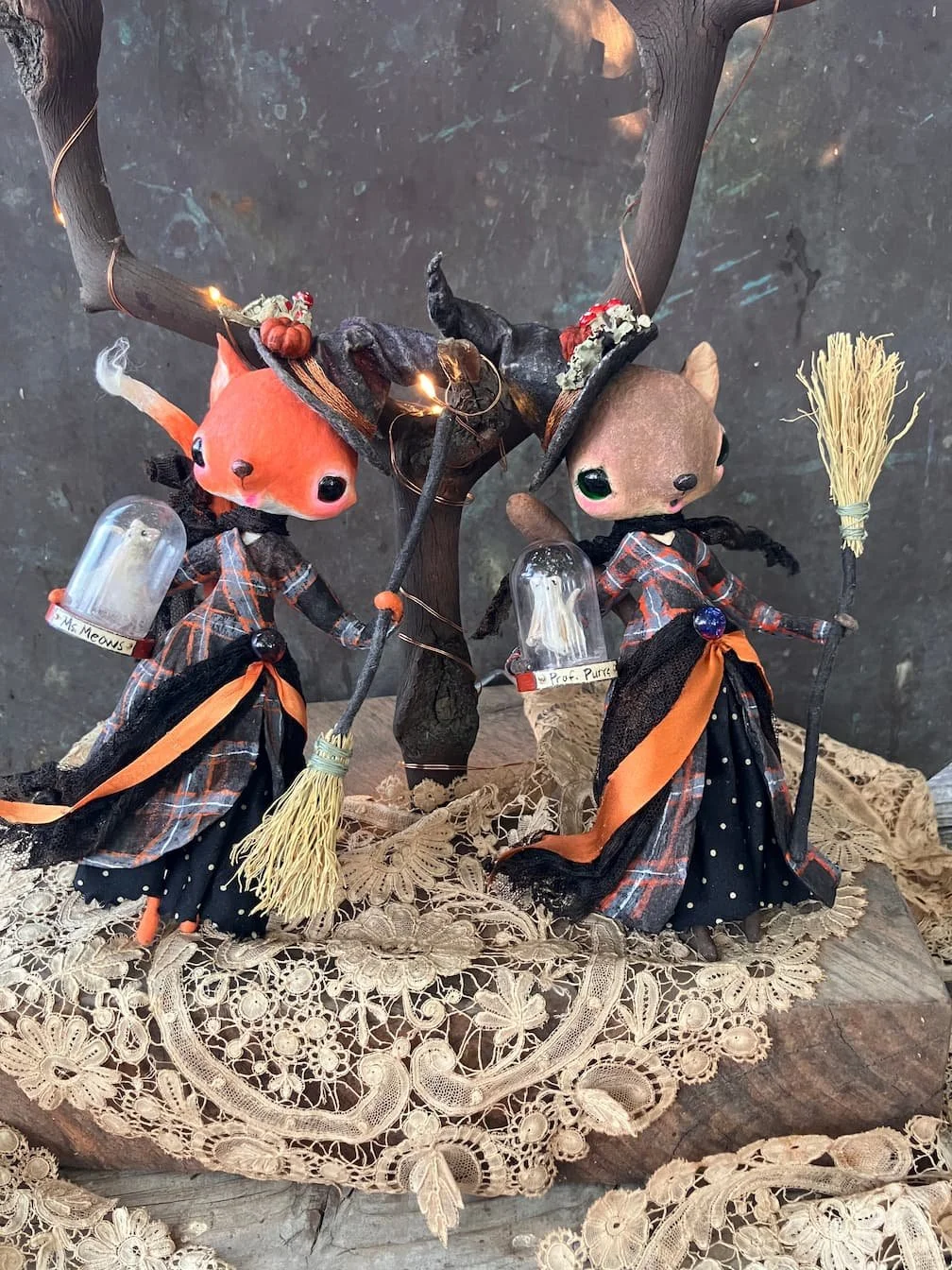 Two spun cotton animal ornaments dressed as witches, standing on a lace doily, holding broomsticks, with a small tree decorated with warm fairy lights behind them.