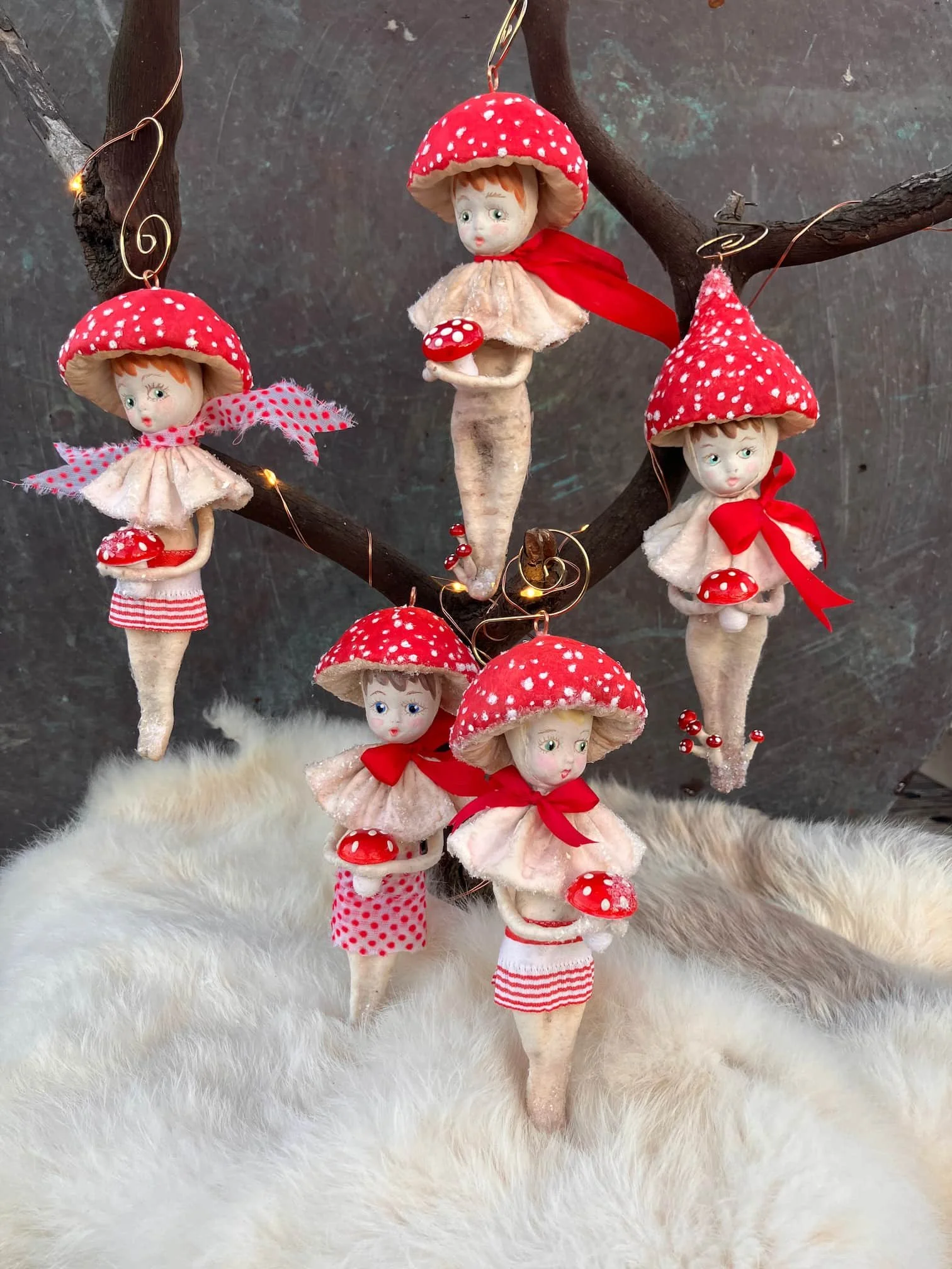 Decorative autumn tree branch with hanging plush dolls dressed like mushrooms, featuring red caps with white spots, beige bodies, and red ribbons, set against a dark background and placed on a white furry surface.