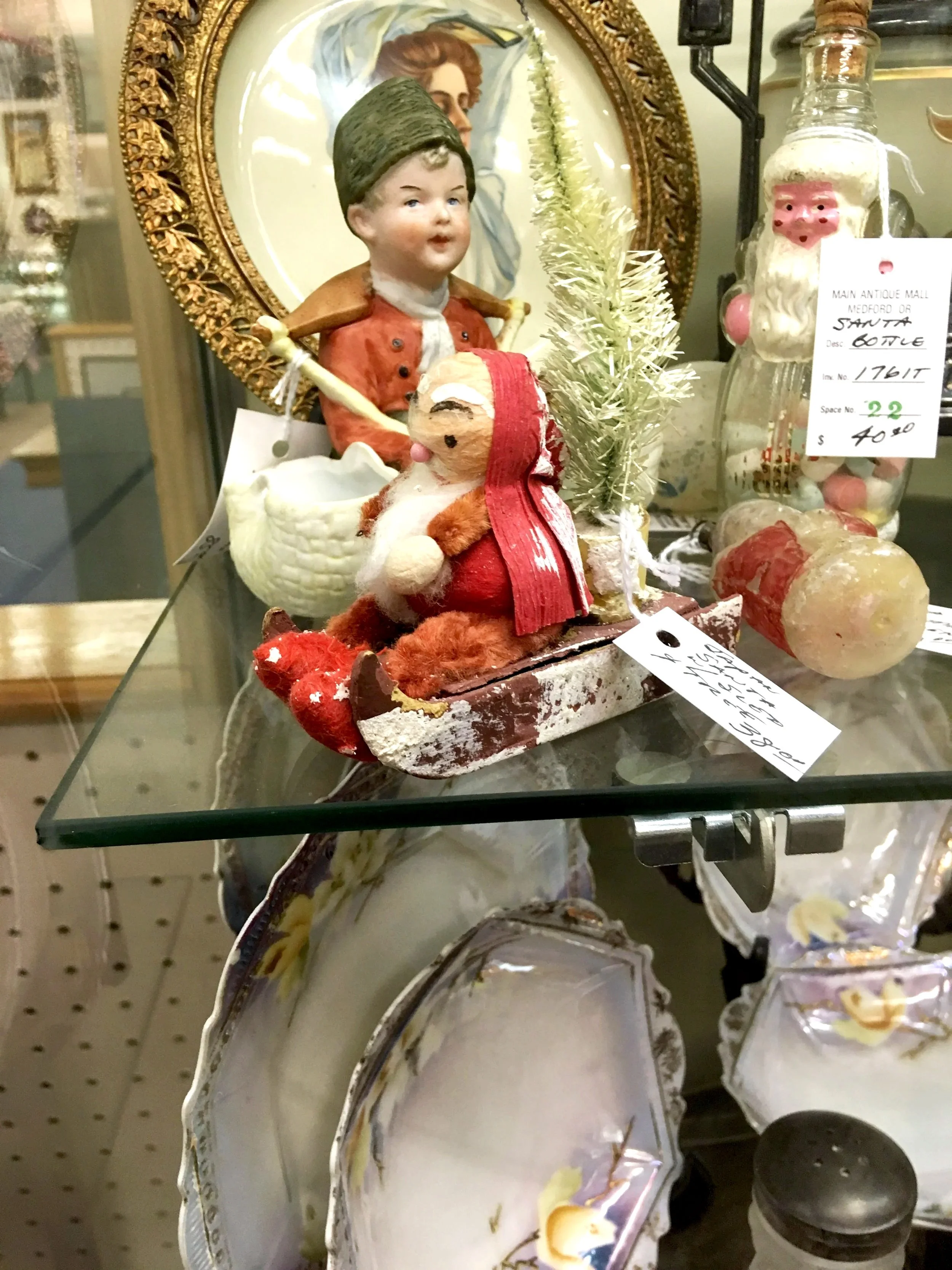 Vintage Christmas decorations including a figurine of a boy holding a stick, a vintage spun cotton Santa, a small artificial pine tree, and a Santa Claus figure in a glass container, displayed on a glass shelf.