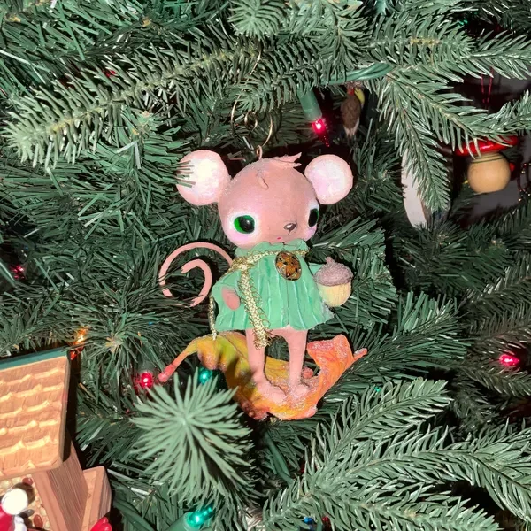 A plush mouse ornament dressed in a green dress with a gold chain and a pin, hanging on a decorated Christmas tree.