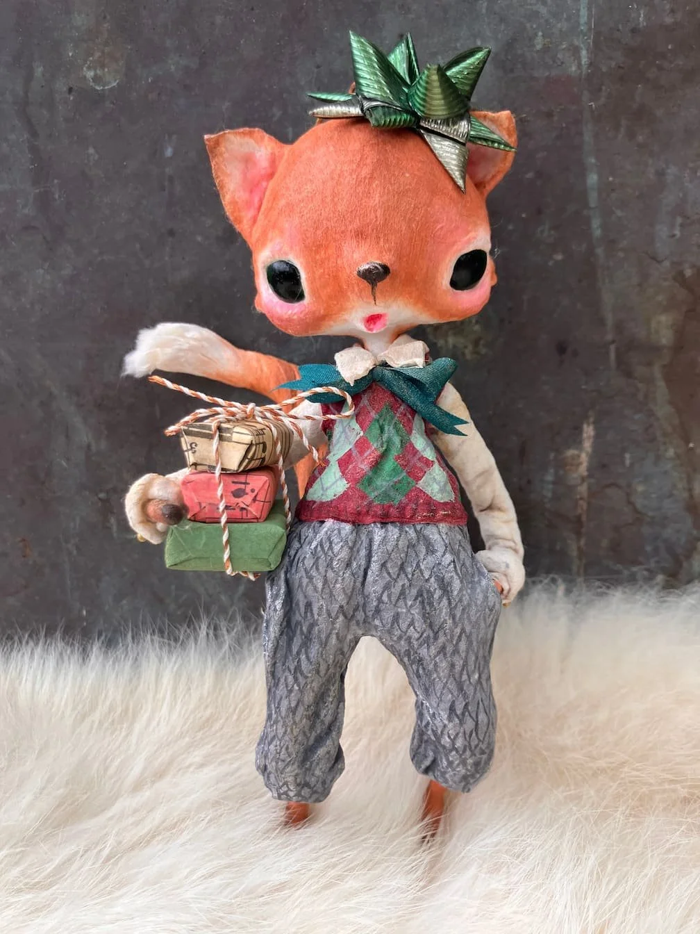 A plush toy of a fox with large black eyes, standing on a white furry surface against a dark background. The fox has a green leaf headpiece, a colorful vest, gray patterned pants, and holds small wrapped gift boxes with twine.