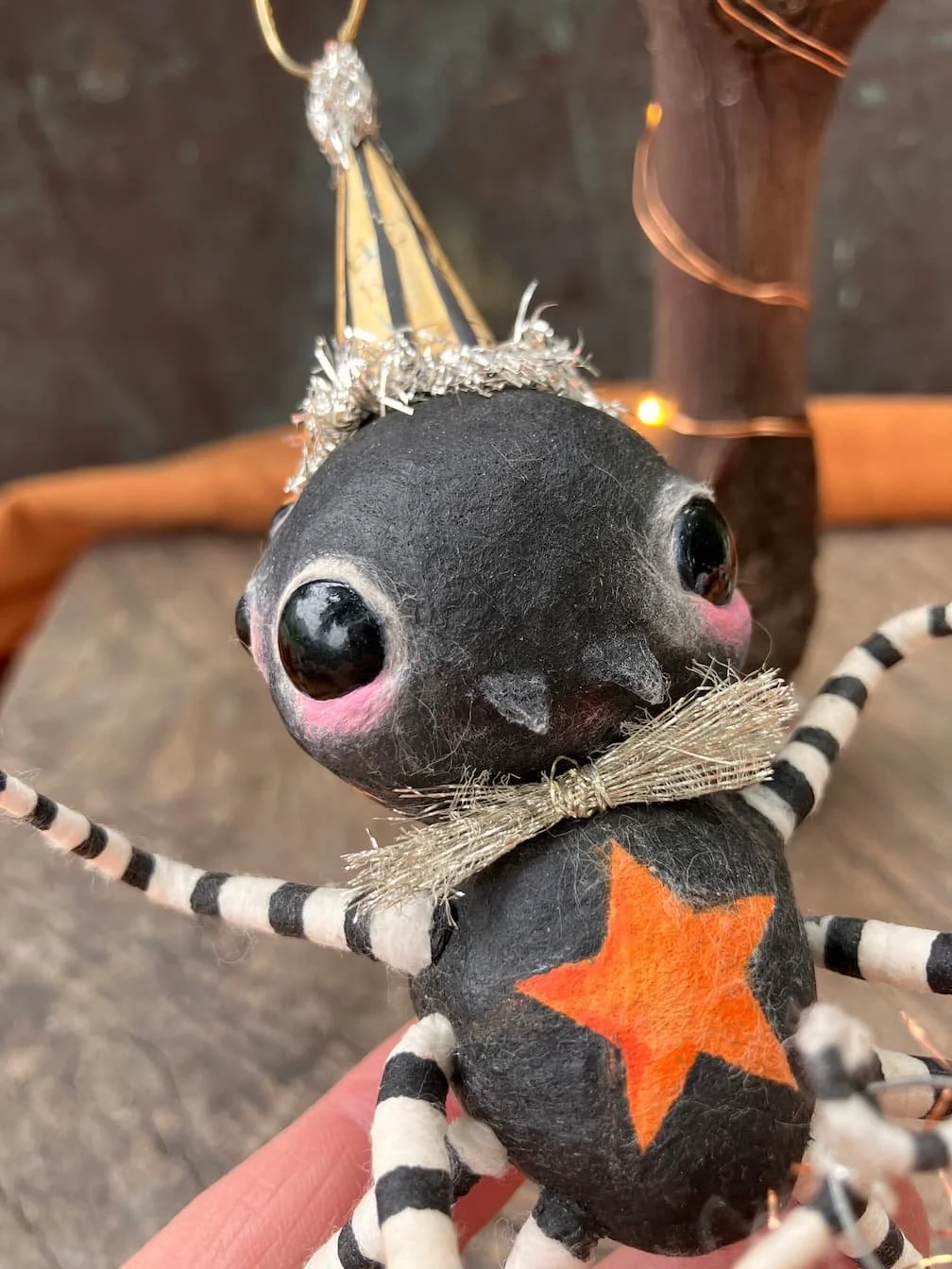 A black Halloween decoration with large eyes, pink blush on the cheeks, wearing a striped black and white outfit, with an orange star on its chest, and a small silver bow tie.