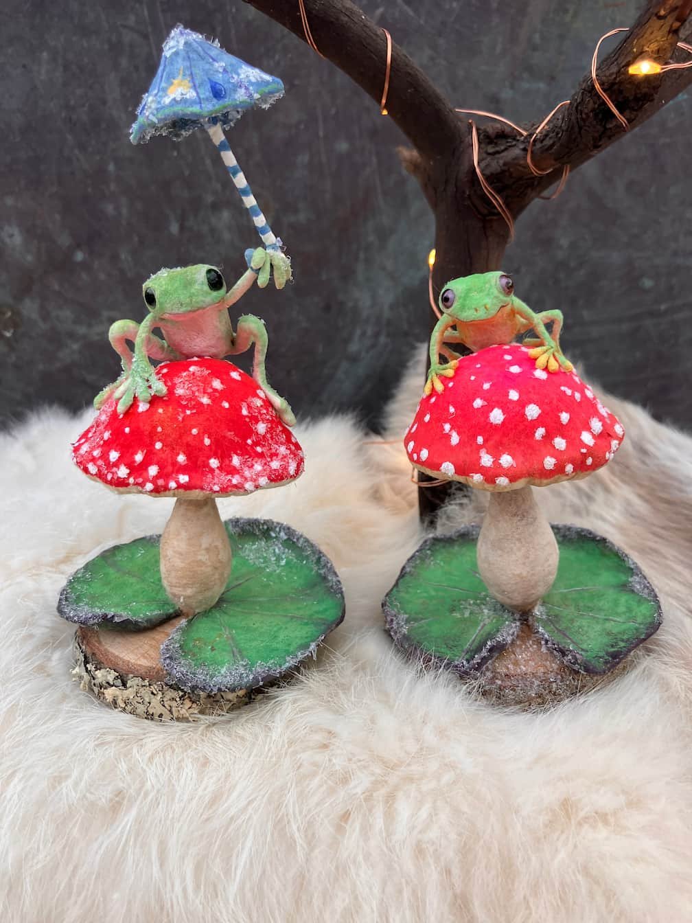 Two decorative frog figurines sitting on red mushroom with white spots, green leaves, and a small wooden base, against a dark background with a branch and string lights.