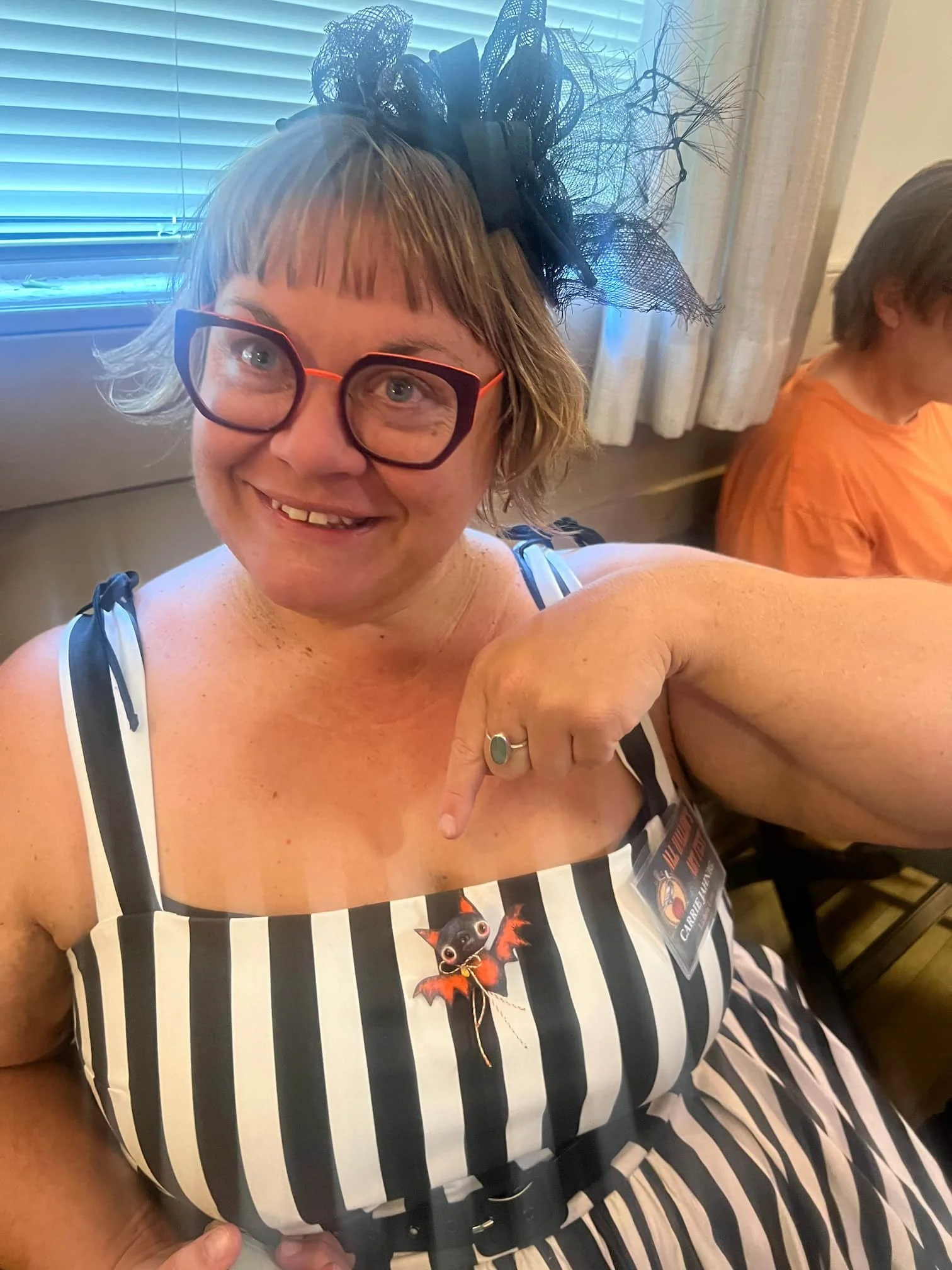 A woman with glasses, a short blonde haircut, and a big black fascinator headpiece, smiling and pointing at her dress which features a cartoon bat and black and white stripes. She is sitting indoors with a window and blinds behind her. Another person with short hair wearing an orange shirt is partially visible in the background.