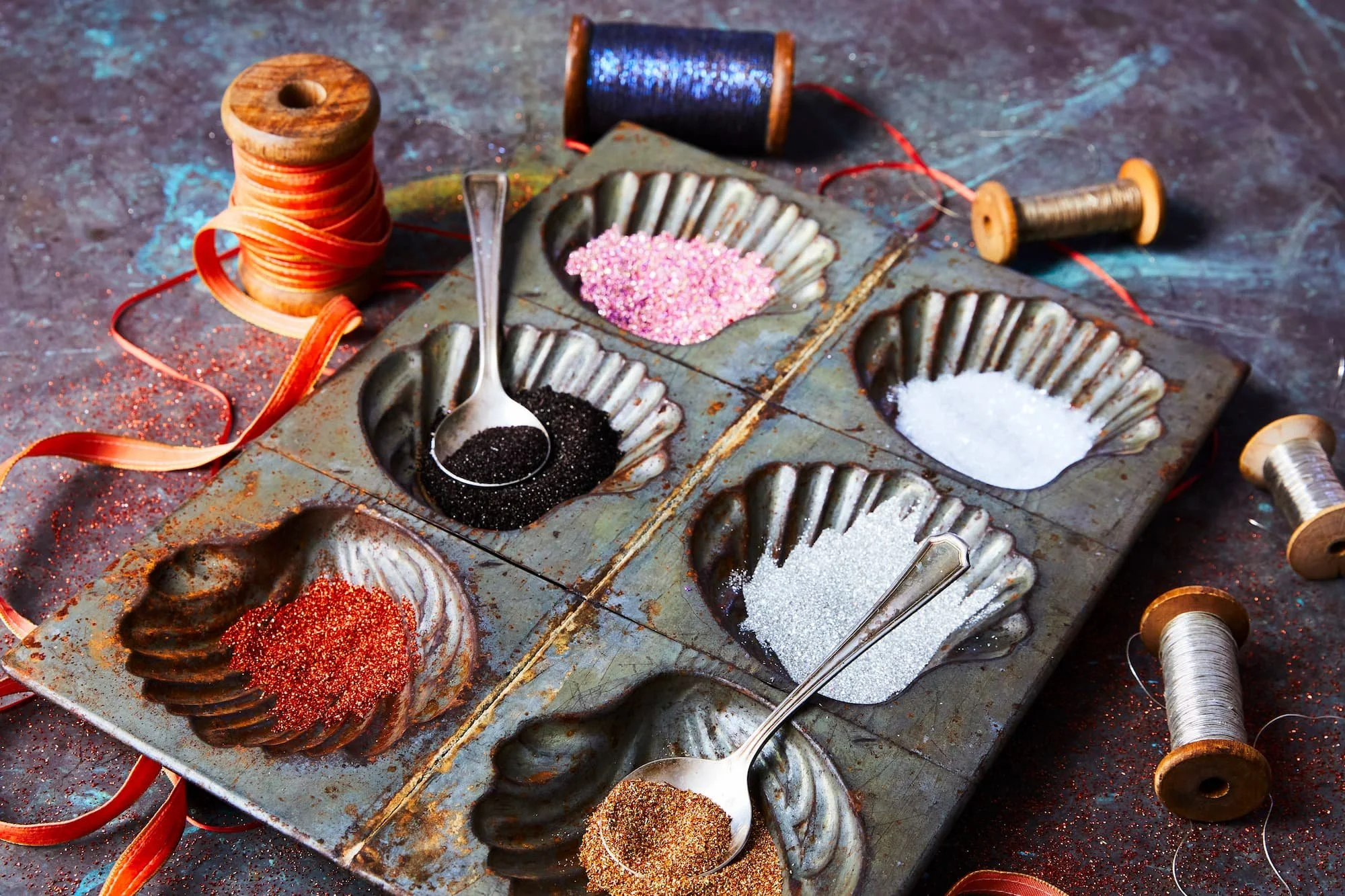 A vintage metal spice rack containing four compartments filled with red, black, pink, and white spices or glitter. Several wooden spools of thread and loose glitter are scattered around, with some spilled onto the dark surface beneath.