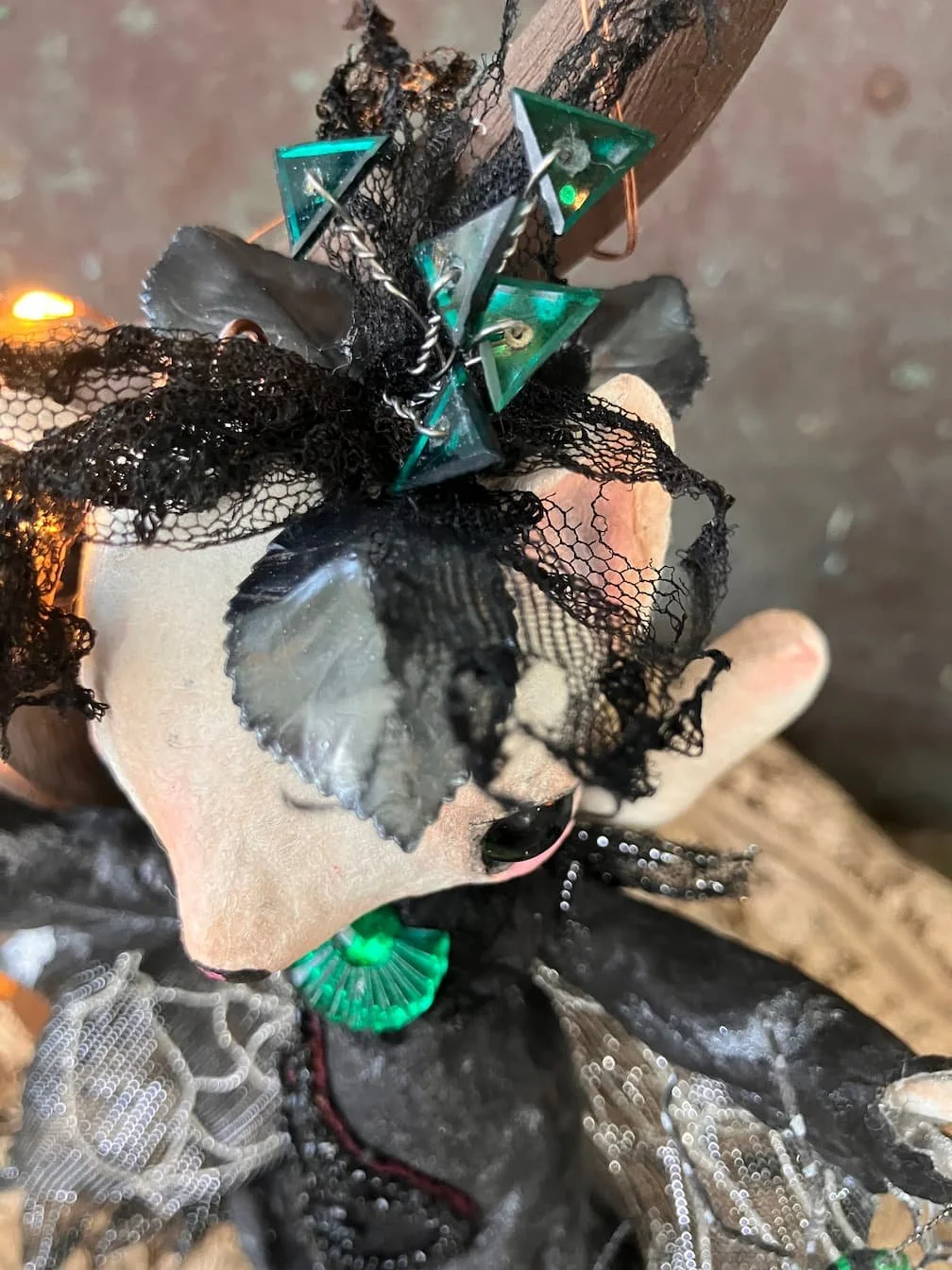 Close-up of a small sculpture featuring black lace, feathers, and green glass or plastic triangles attached to a wooden stick.