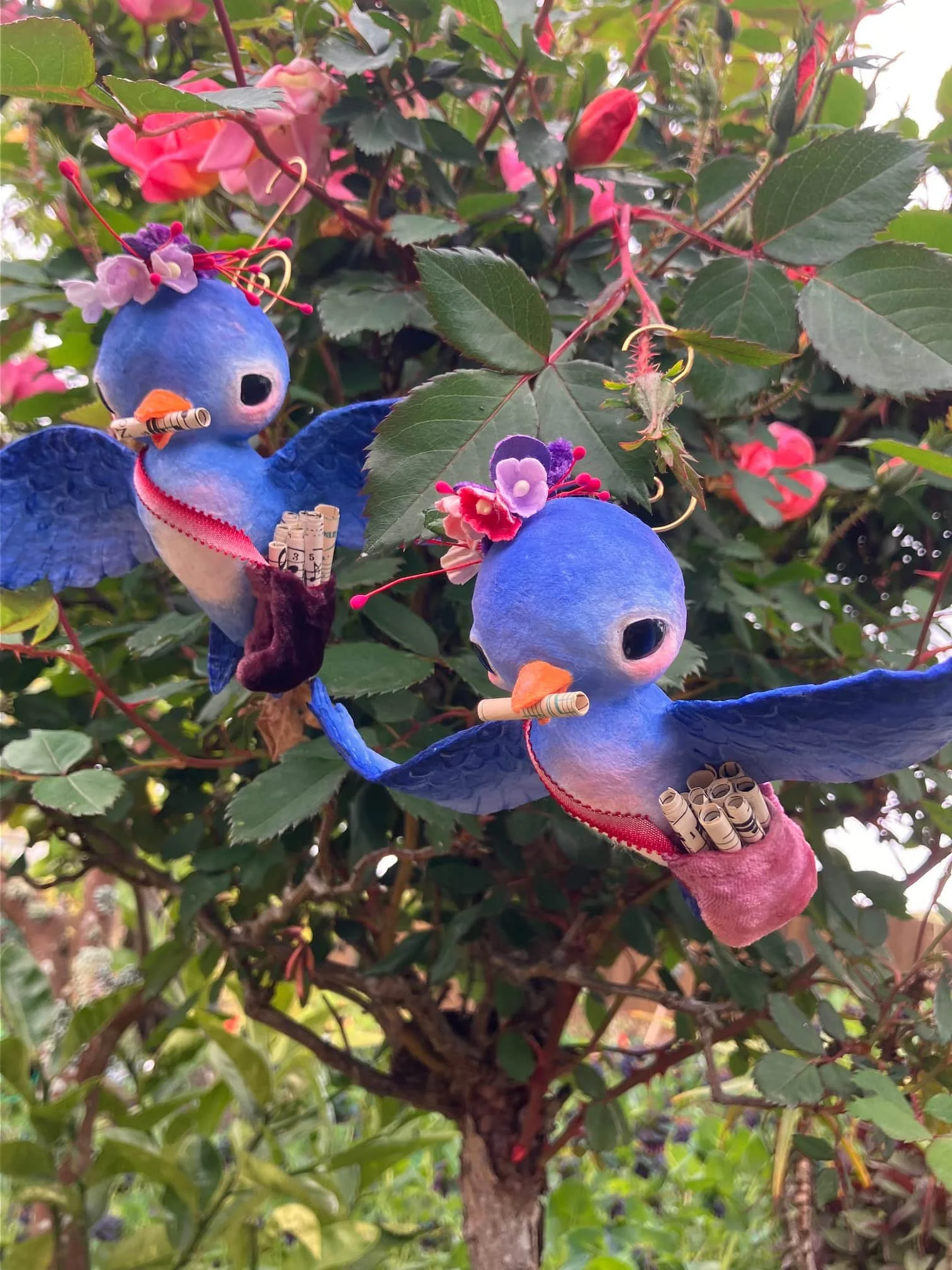 Decorative blue bird ornaments placed on a bush with green leaves and pink flowers.
