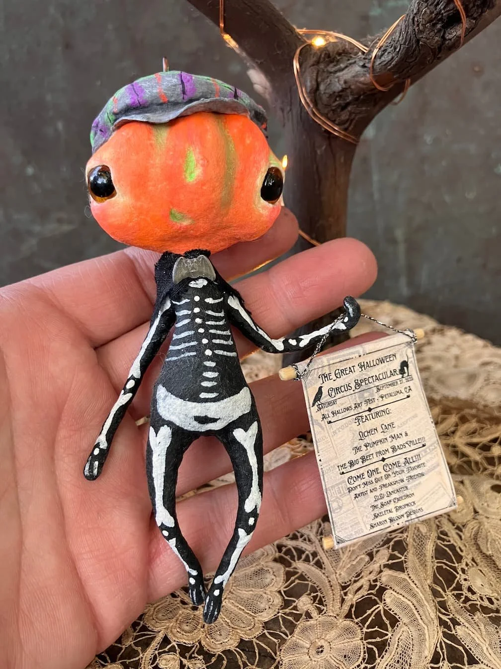 A sculpture with an orange pumpkin head, large black eyes, and a painted skeleton body, wearing a colorful cap, holding a sign for a Halloween event, with a wooden tree and string lights in the background.