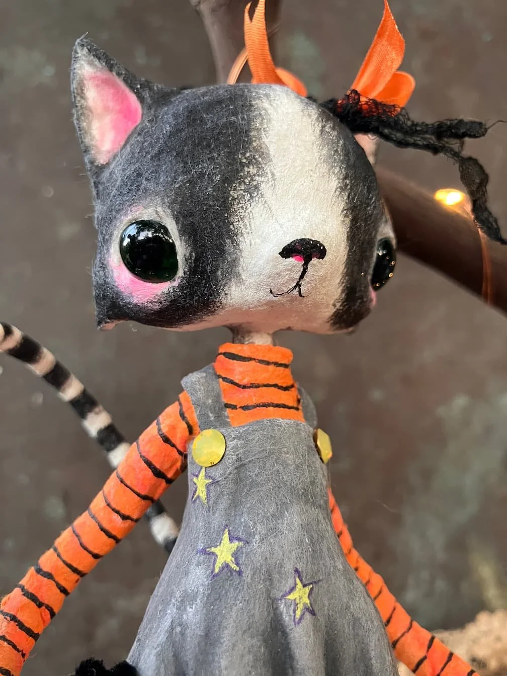 A close-up of a handmade cat doll with black and white fur, large shiny black eyes, pink cheeks, and a small black nose. The doll wears an orange and black striped shirt, a gray dress decorated with purple stars, and has orange and black ribbons on i