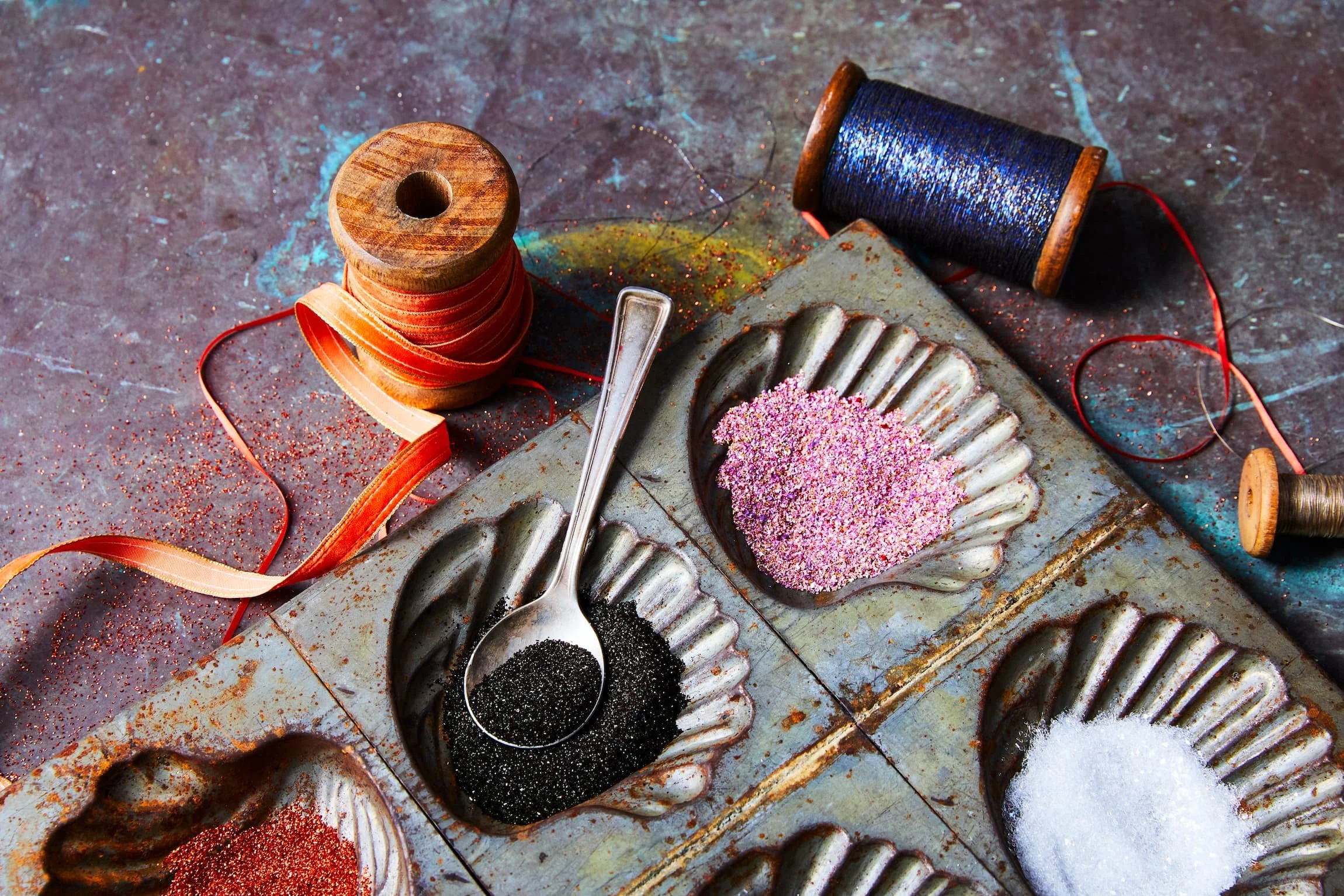 Spools of thread, a spoon with black glitter, pink glitter, and a rusty baking mold on a colorful, textured surface.