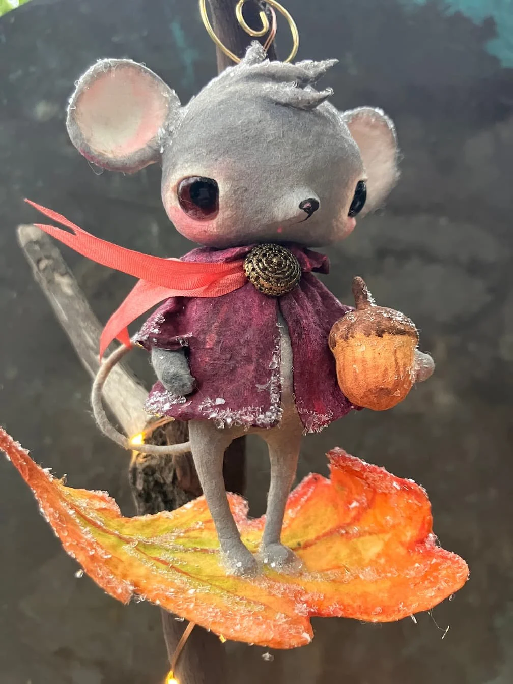 A craft mouse with large black eyes, gray fur, and pink cheeks, wearing a red cape with a gold brooch and holding a small orange pumpkin, standing on a colorful autumn leaf with a dark background.