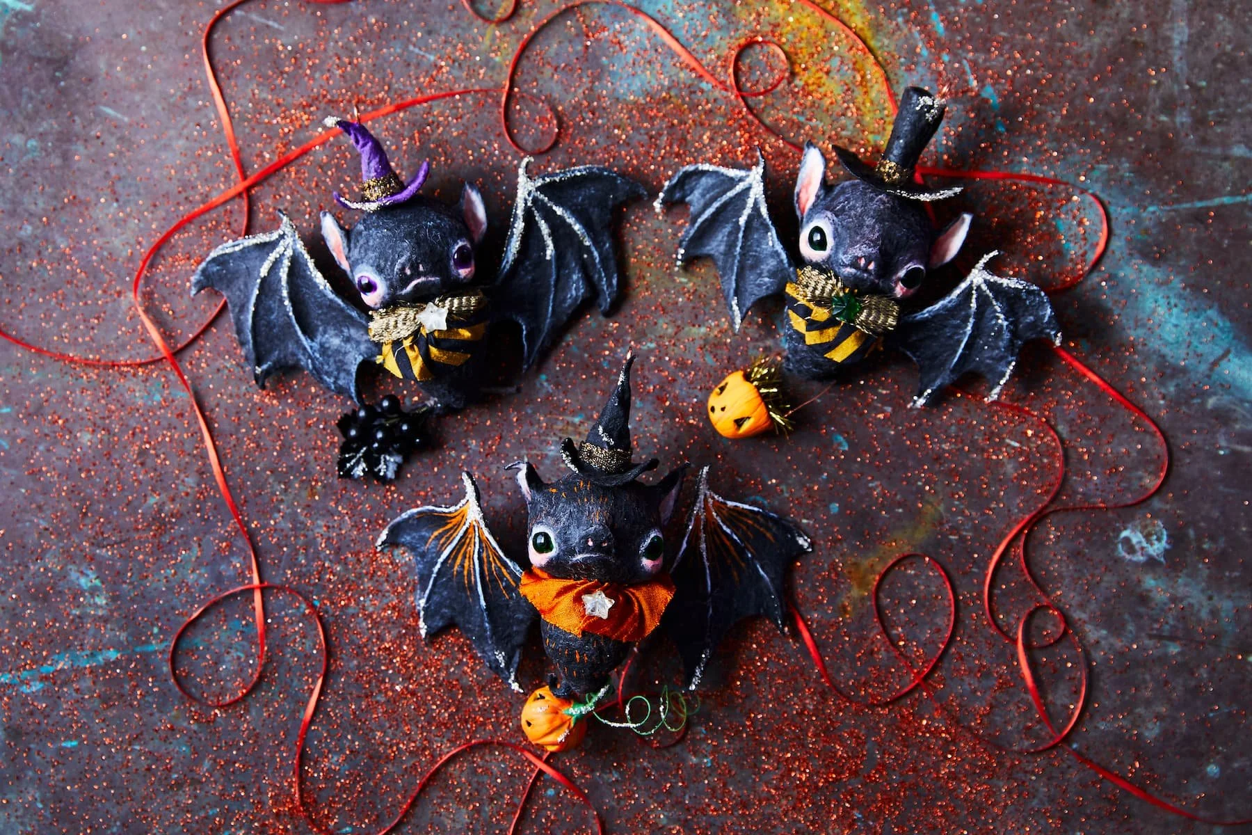 Decorative Halloween arrangement with three small black bat and cat figurines dressed in costumes, surrounded by red string, glitter, and small pumpkin and black berry ornaments on a glittery surface.