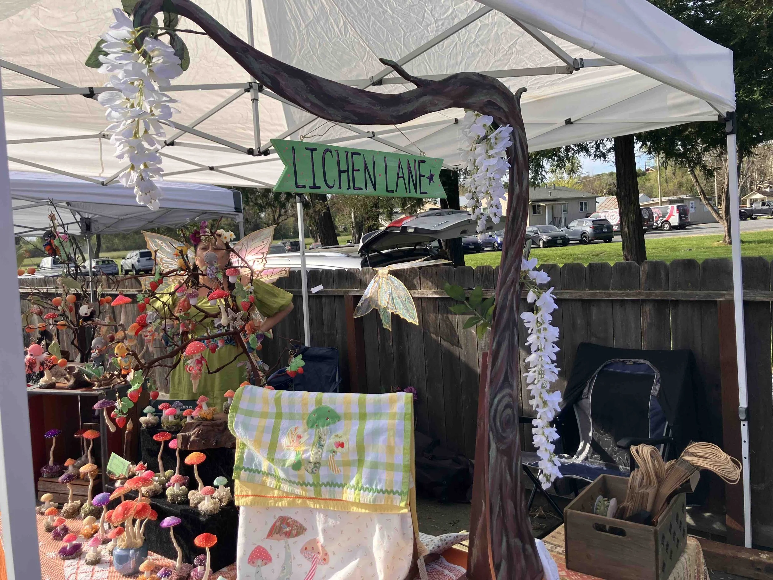A booth at an outdoor market with a handmade sign labeled 'Lichen Lane' hanging from a tree branch. The booth displays colorful felt mushrooms, fairy wings, and decorative items, with a table covered by a checkered cloth and a striped towel.