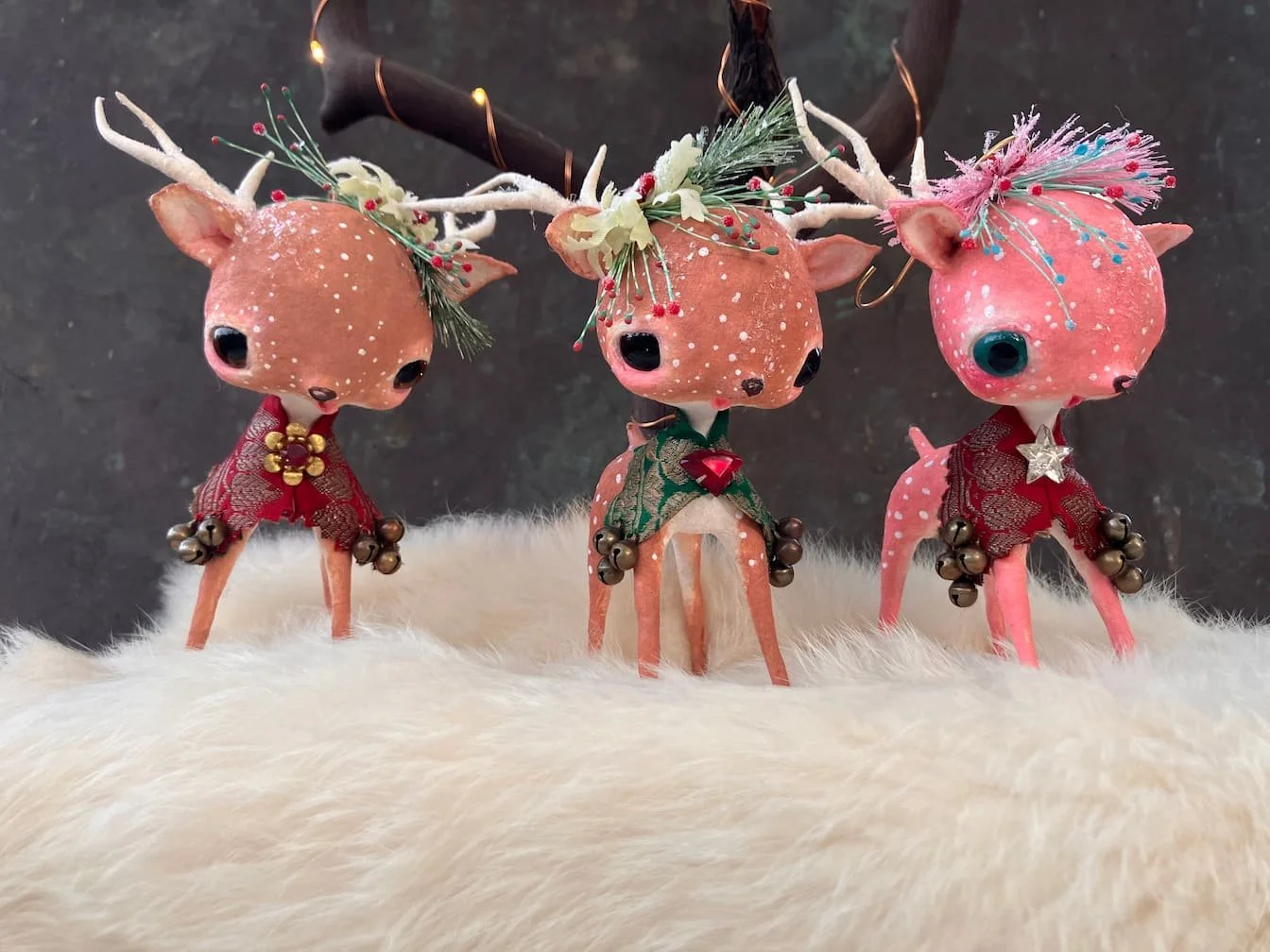 Three decorative reindeer figurines with large eyes and antlers, decorated with Christmas accessories and bells, standing on a white furry surface against a dark background.