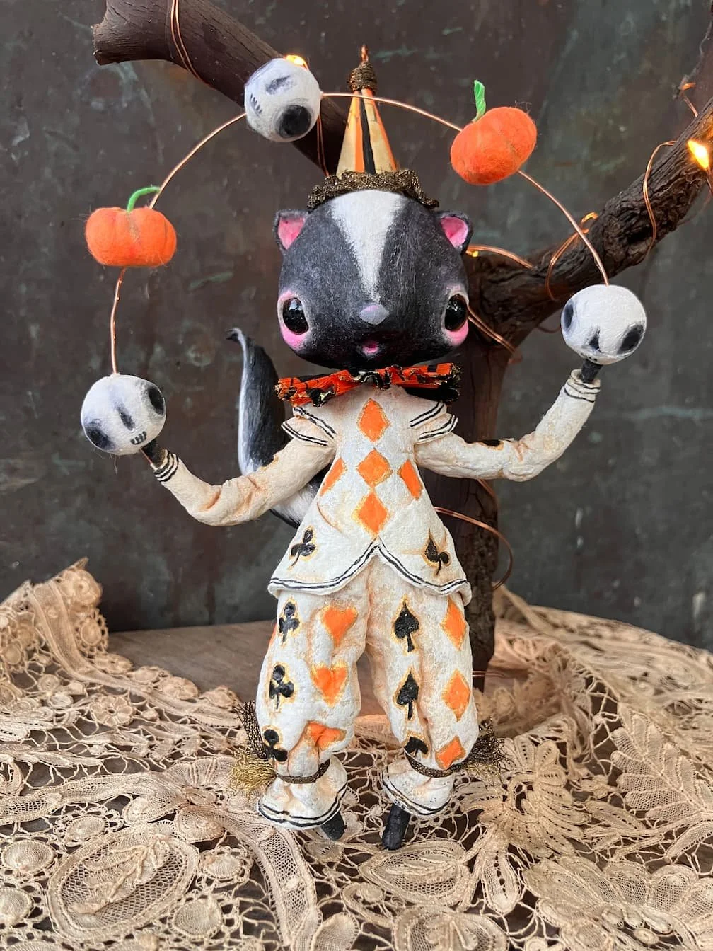 A cute plush toy dressed in a card suit-themed outfit, standing in front of a decorated tree with pumpkin and skull ornaments, on a lace tablecloth.