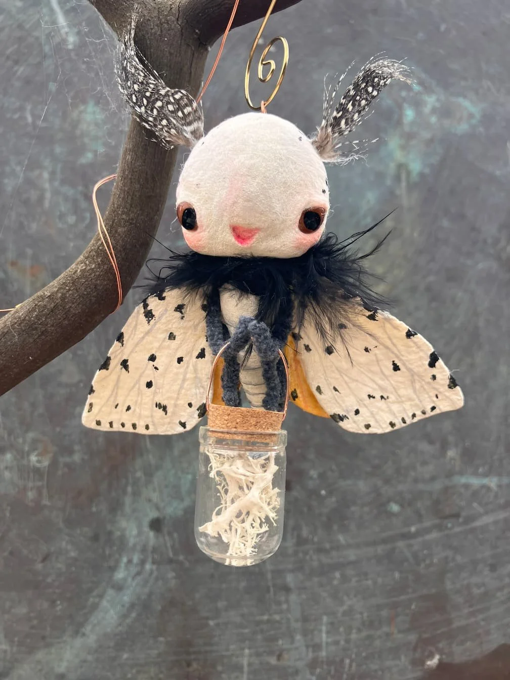 A plush toy resembling a butterfly with a white face, black eyes, pink cheeks, and a small pink mouth. It has feathered antennae and a black ruffled collar. Its wings are white with black spots. The toy hangs from a tree branch with a small glass jar