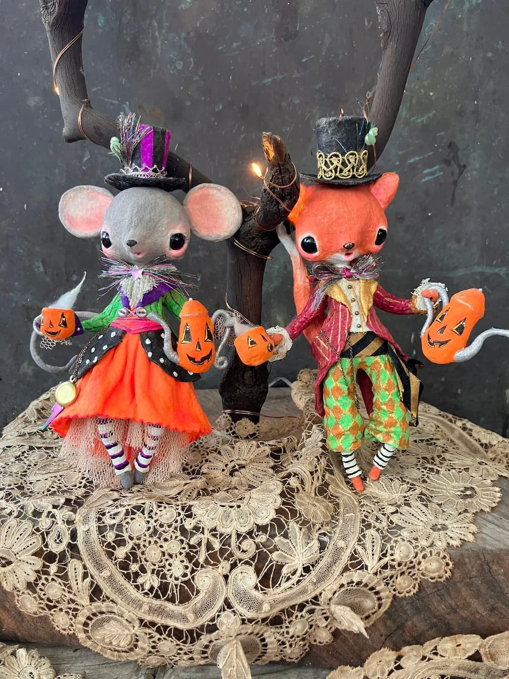 Two Halloween-themed animal figurines, a mouse and a fox, dressed in colorful costumes. They are standing on a lace cloth near a small, decorative tree with lights. Both animals are holding small pumpkin buckets for trick-or-treating and are wearing 