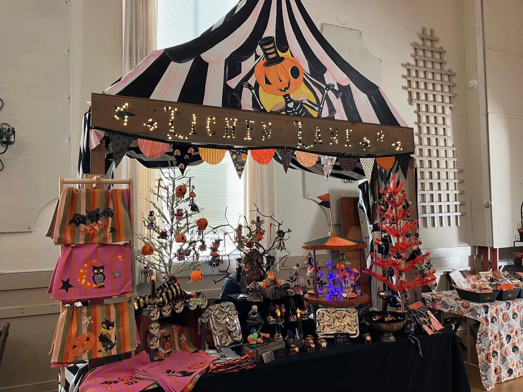 Indoor market stall decorated with Halloween-themed items, including pumpkins, skeletons, and bats, as well as a sign with lights saying 'Luchan Lane.' A pumpkin with a top hat is painted on a large backdrop.