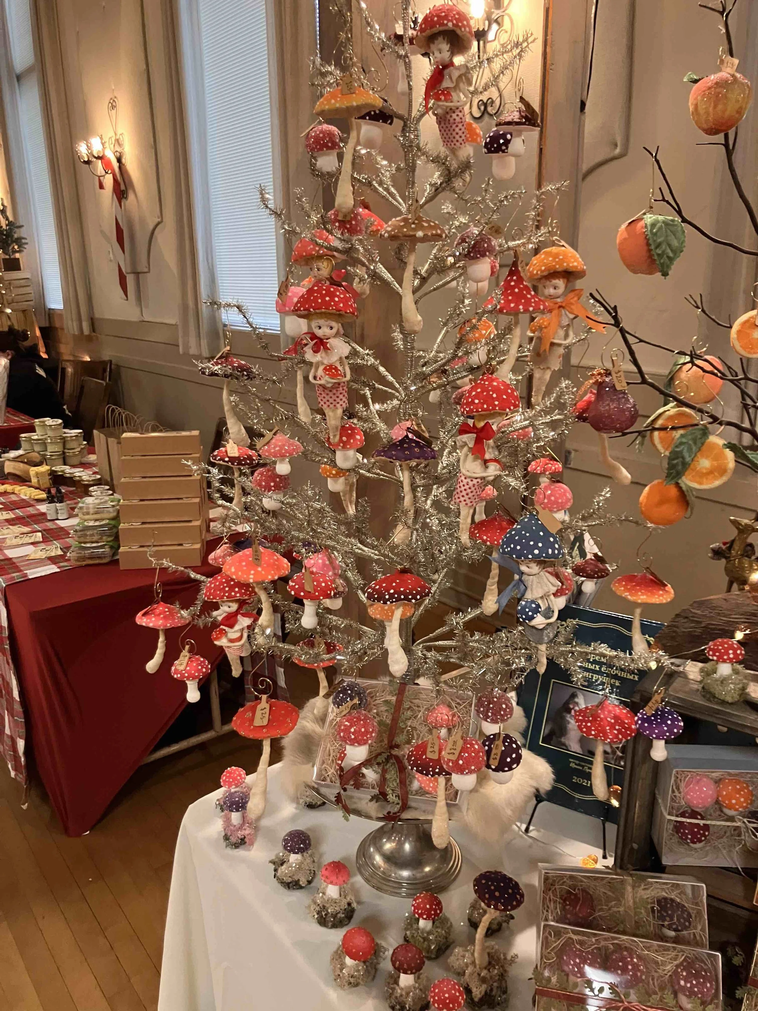 A table displays various mushroom-themed ornaments, with several doll-like figurines dressed as mushrooms hanging from a silver-colored tree and others placed on the table.