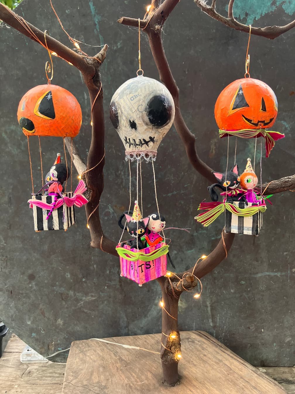 Decorative Halloween tree with hanging paper lanterns shaped like pumpkins and a skull, along with plush black cats and other Halloween-themed plush toys in small boxes, all illuminated by string lights.
