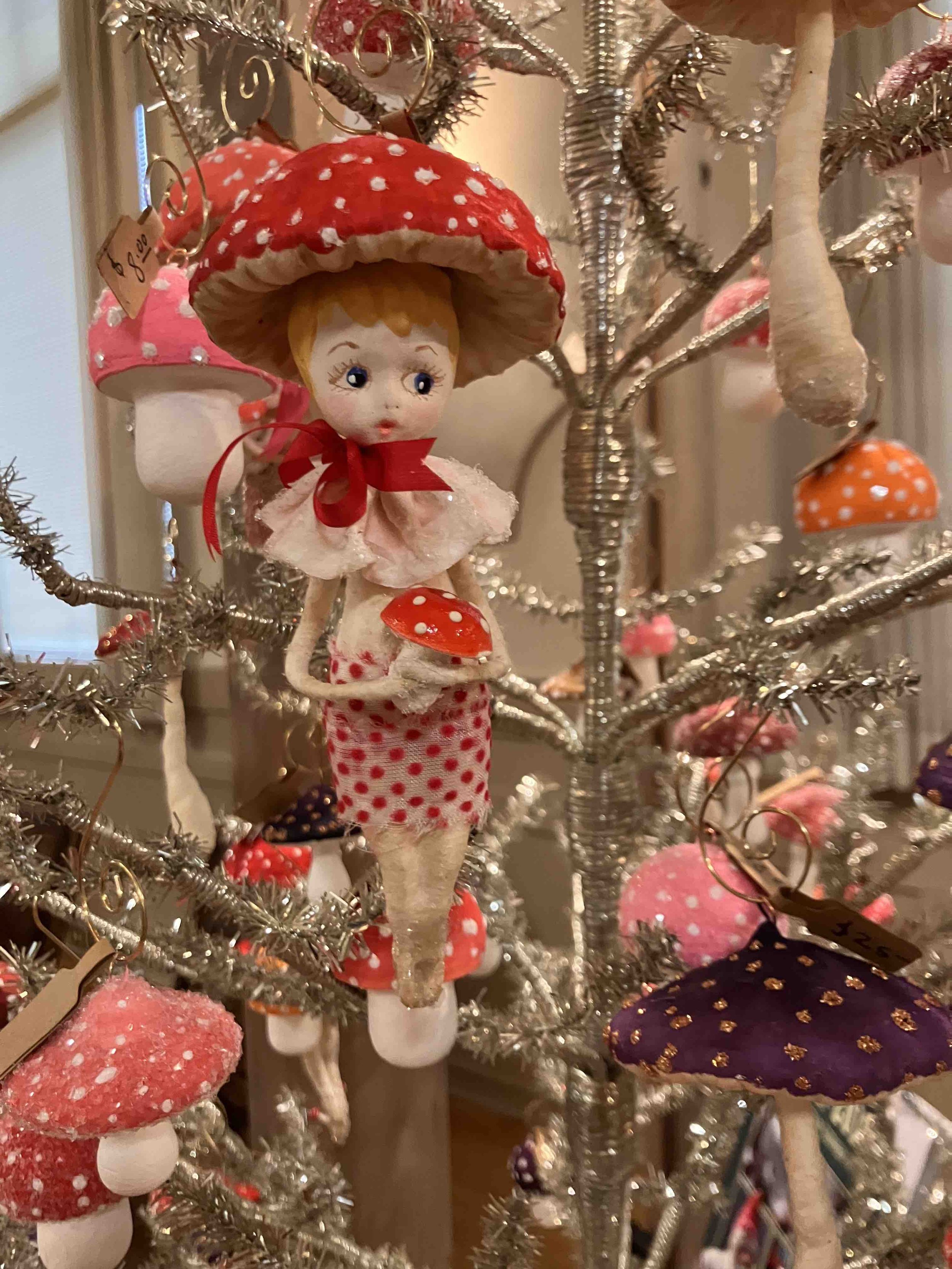 Close-up of a Christmas tree decorated with whimsical mushroom ornaments, including a doll-like figure with a mushroom cap as a hat, surrounded by various pink, red, and purple mushroom decorations, and silver tinsel.