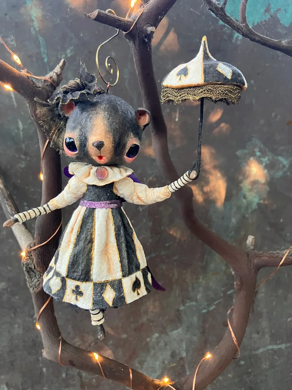 A doll of a mouse dressed in a black and gold queen or princess costume with a dress, cape, and striped stockings. The doll has big eyes, a nose, and is holding a small parasol. It is hanging on a twisted branch decorated with fairy lights, with a da
