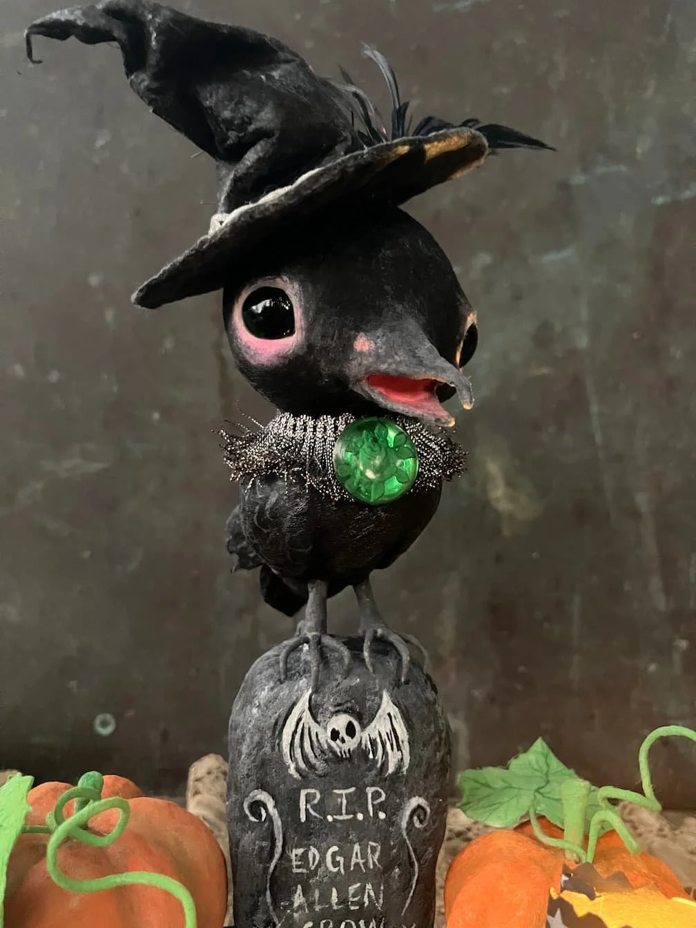 A black bird figurine with large eyes, wearing a witch's hat and a sparkly collar featuring a green gem, standing on a tombstone with a skull and crossbones inscription reading 'R.I.P. Edgar Allen,' surrounded by orange pumpkins and green leaves.