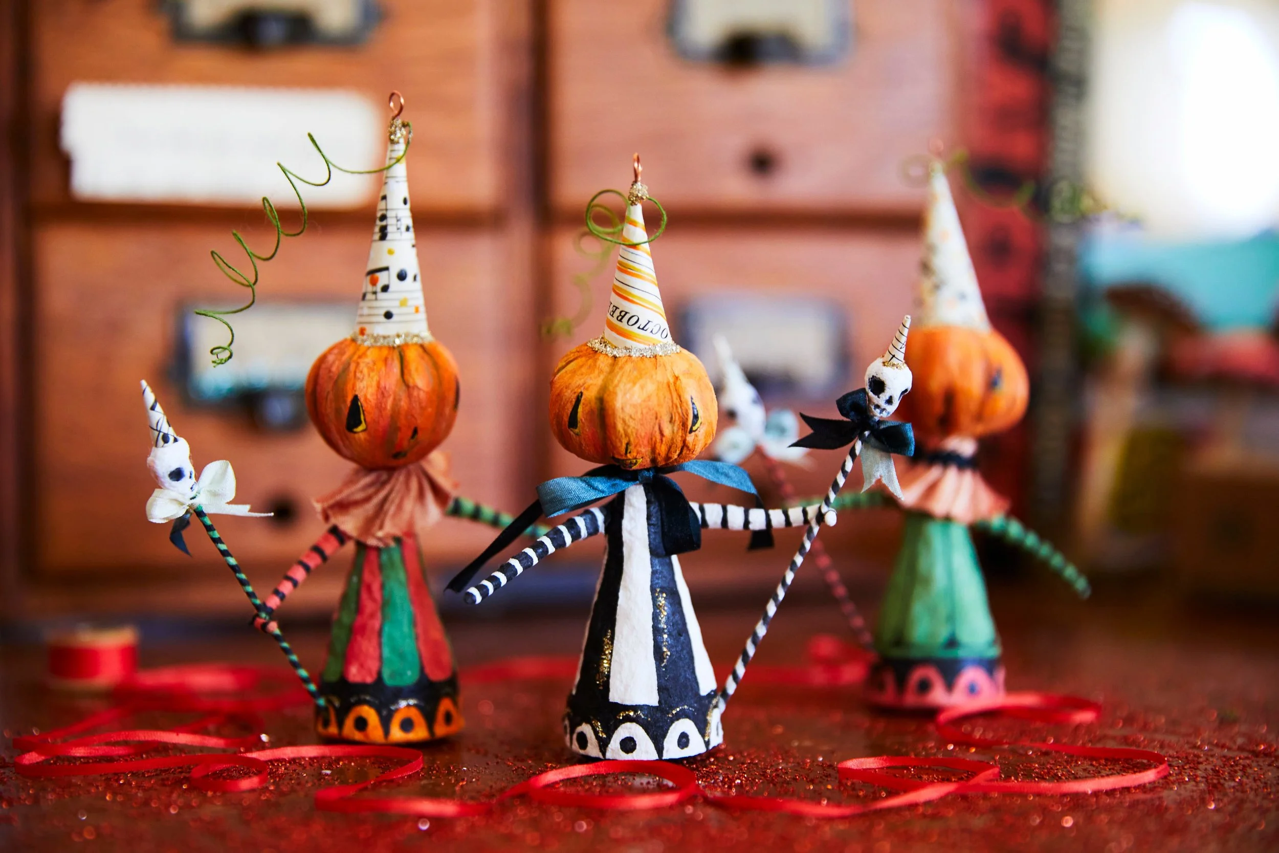 Decorative Halloween figurines with pumpkin heads wearing colorful cone hats and skeleton arms, set on a red glittery surface with scattered red spirals.