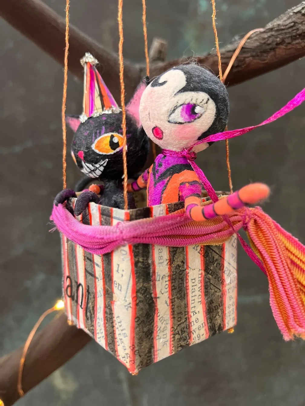 Handmade plush dolls of a cat and a woman with pink hair in a colorful fabric basket hanging from a branch with string lights.
