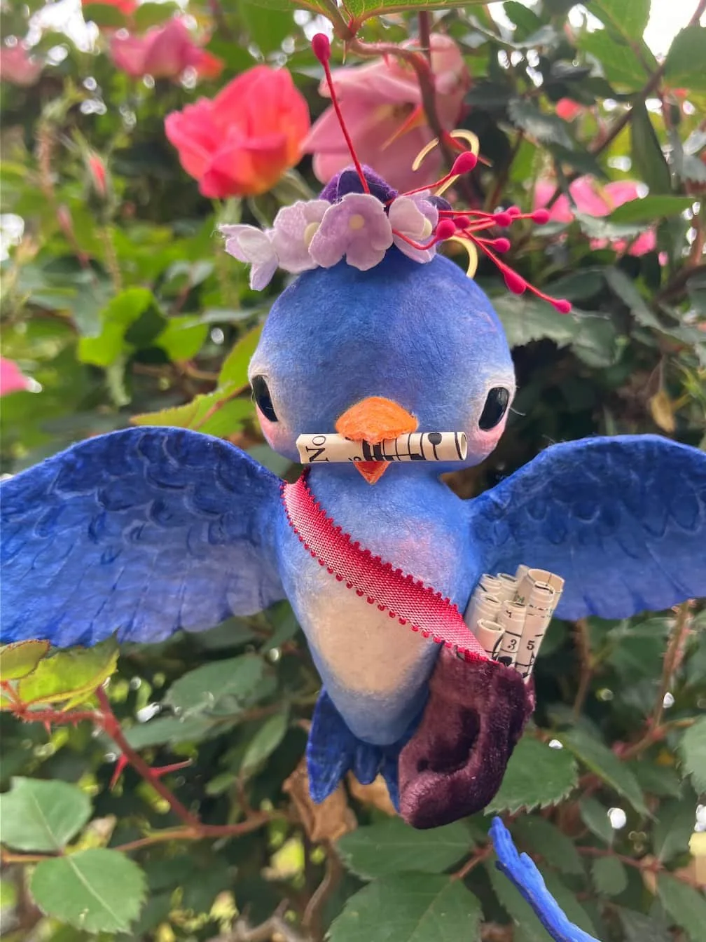 A plush blue bird with wings spread, decorated with pink flowers and wearing a red ribbon, holding rolled-up papers in its beak and paw, surrounded by green leaves and pink flowers.