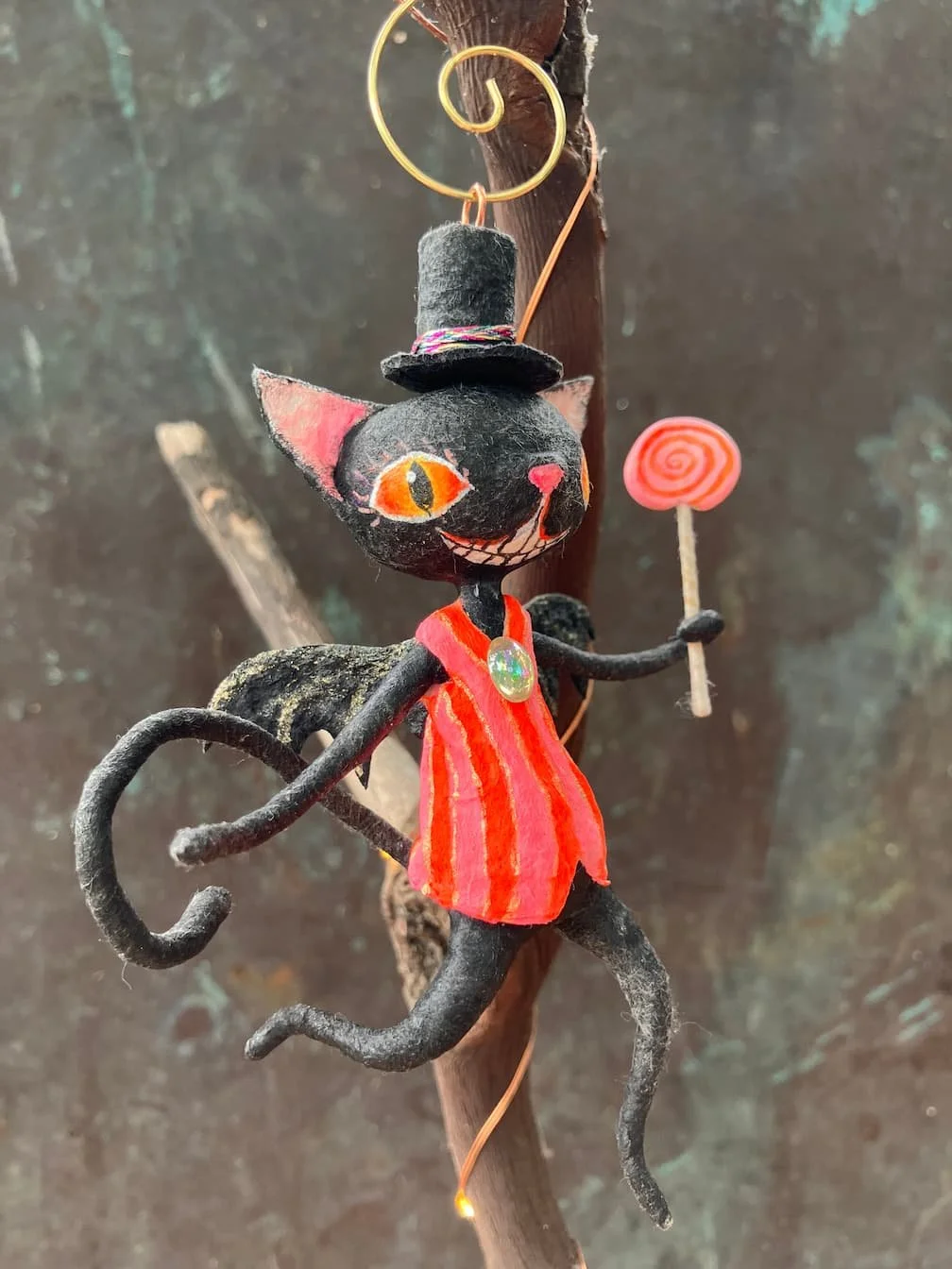 A whimsical black cat figurine with orange eyes, wearing a striped red and orange dress and a small black top hat. The cat is holding a red lollipop and has a curled tail. It is hanging from a stick attached to a wooden branch with a copper wire.