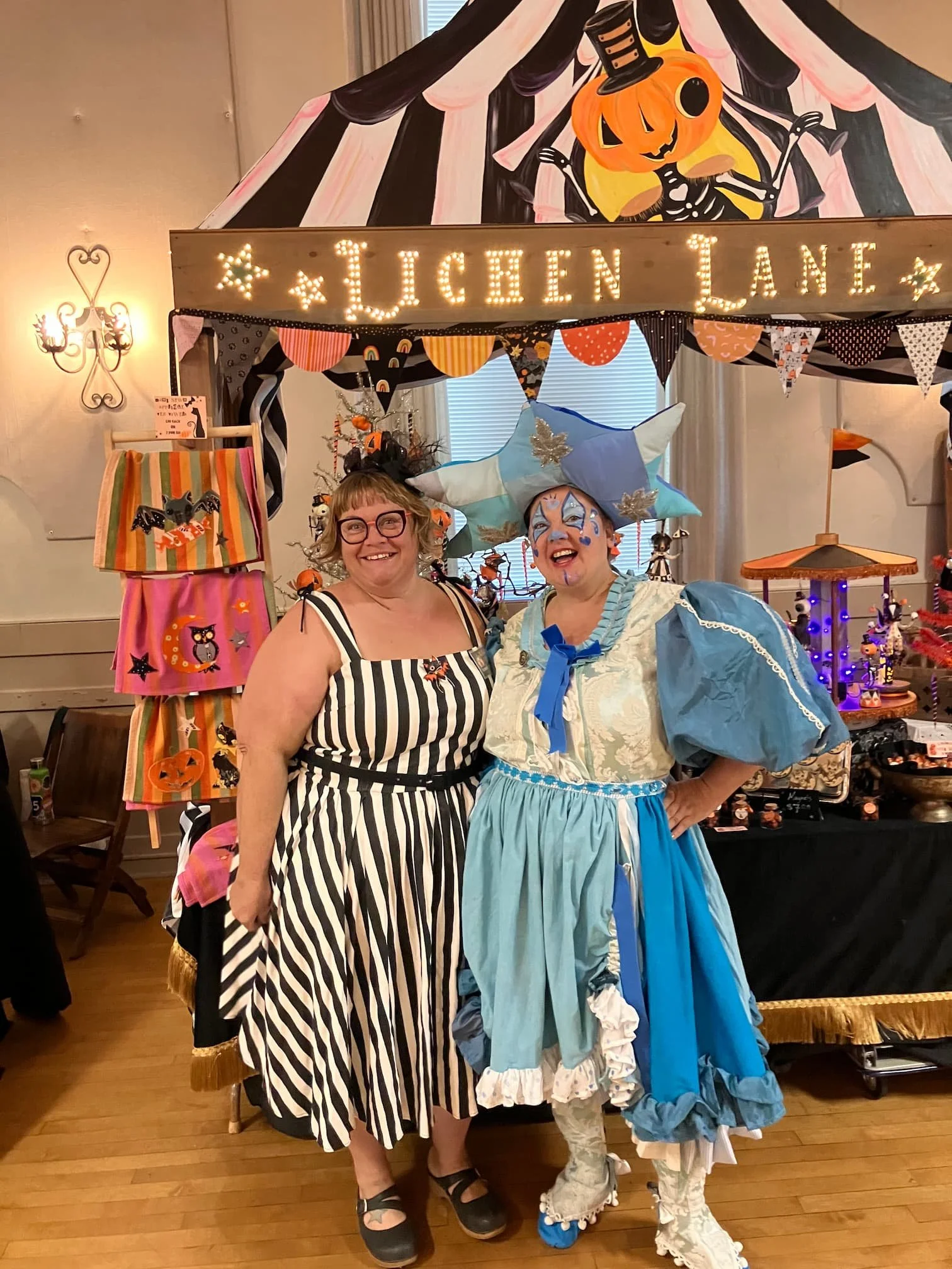 Two women dressed in Halloween costumes standing in front of a decorated booth with a sign that says 'LUNCH LINE' and a Halloween-themed backdrop with a pumpkin skeleton. One woman is dressed as a clown or fairy tale character with a large ruffled dress and cross jewelry, and the other as a witch with glasses and a striped dress featuring Halloween motifs.