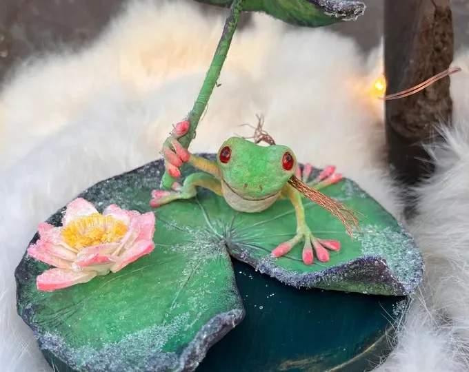 Decorative frog figurine sitting on a lily pad with a pink flower, surrounded by fluffy white material and fairy lights.