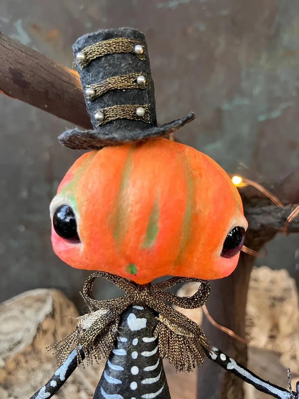 Decorative pumpkin with a small, black, fuzzy hat with gold embellishments, a skeleton-patterned dress, and a polka-dot bow, set against a rustic background.