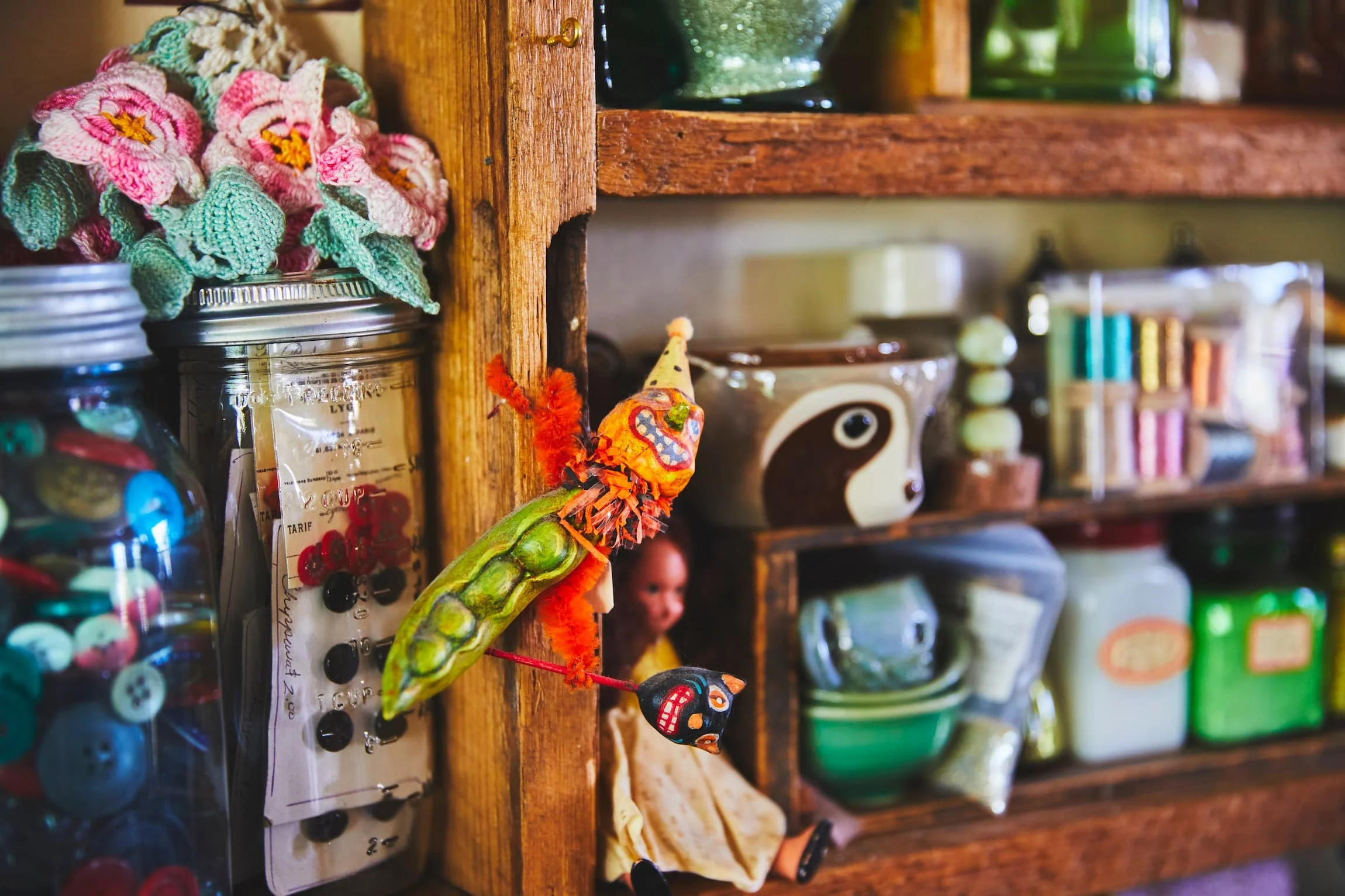 A wooden shelf holding various glass jars and colorful knickknacks, including plush toys and decorative items, with a crocheted flower decoration on the left.