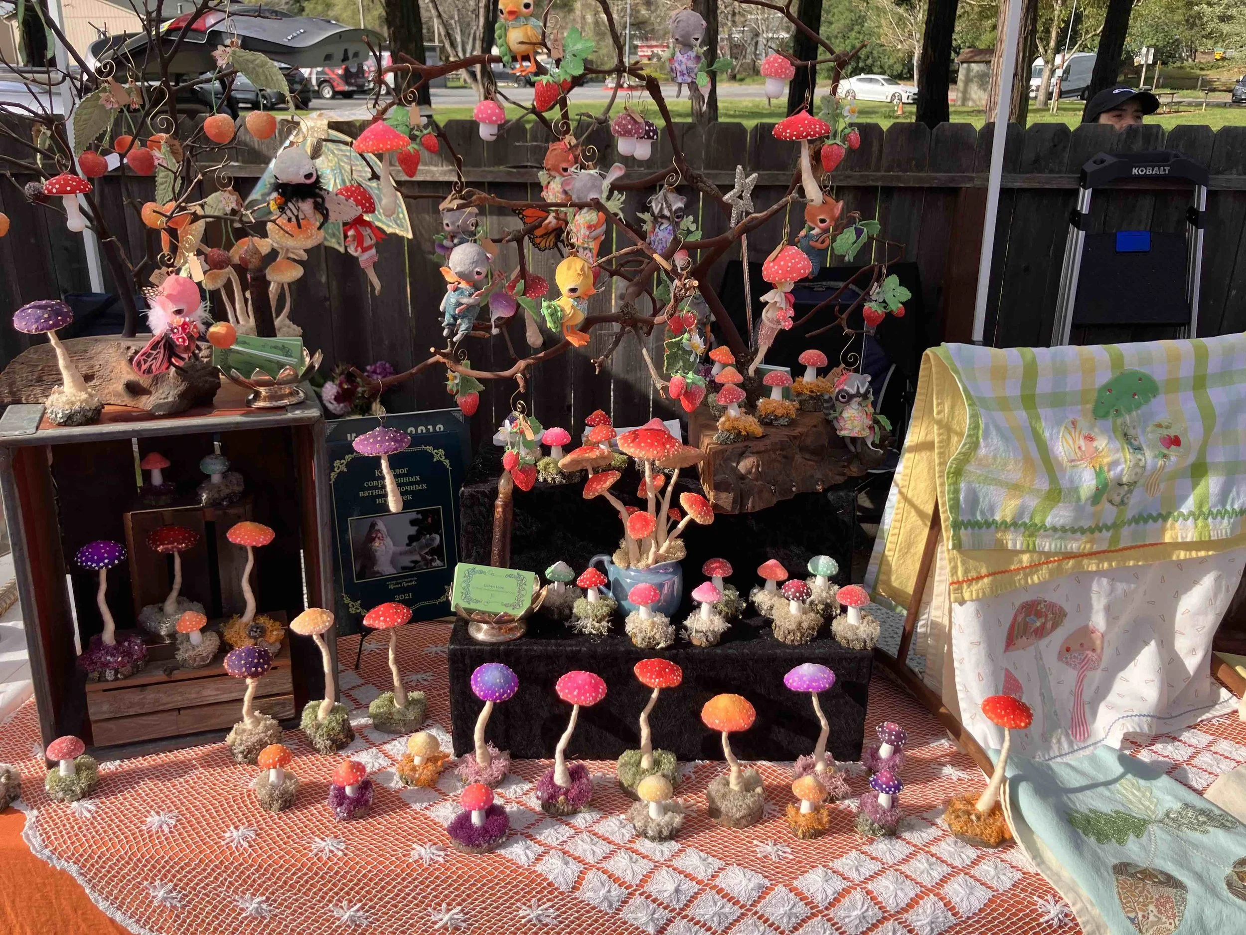 Colorful handcrafted mushroom and fairy ornaments on display at an outdoor market stall, with a wooden fence and cars parked in the background.