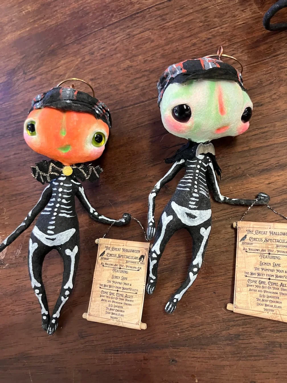 Two Halloween-themed ornaments with skeleton bodies and large heads, one orange and one green, dressed in witch costumes, displayed on a wooden surface with event programs.