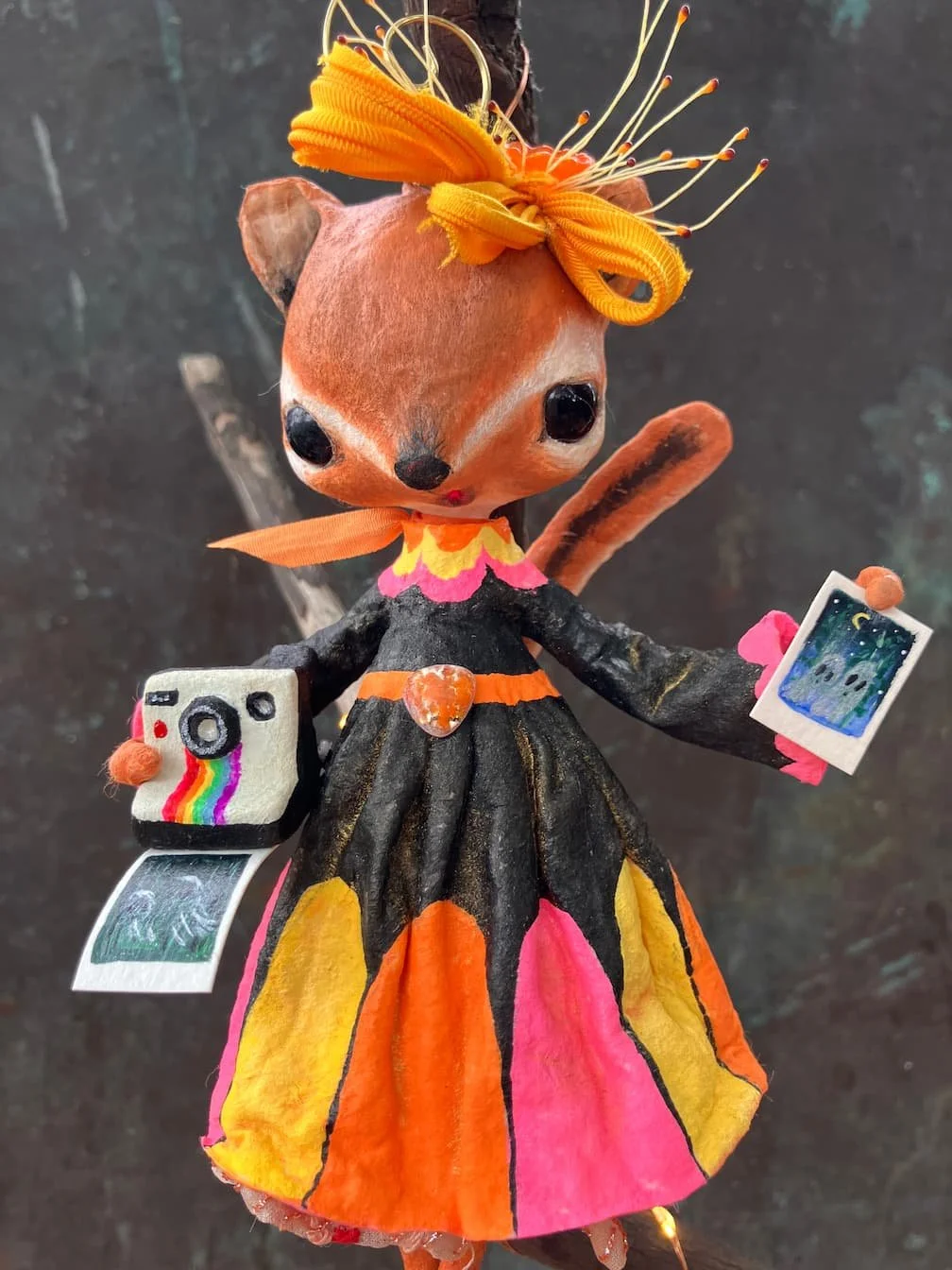 A handcrafted doll of a fox dressed as a photographer, wearing a colorful dress with pink, orange, yellow, and black sections. The doll has large black eyes, a small nose, and a tail. It has an orange ribbon around its neck and another on its waist. 