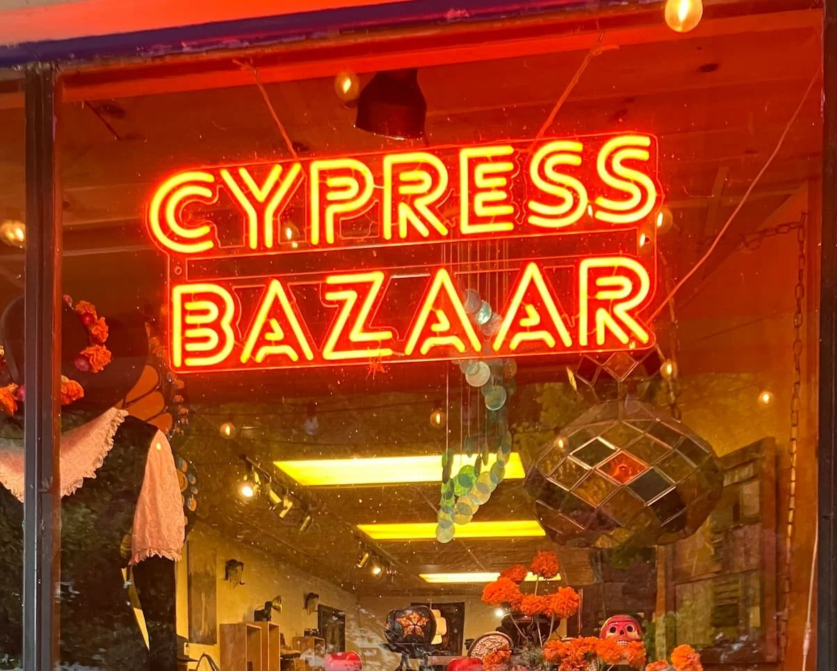 Neon sign reading 'Cypress Bazaar' in bright red letters, with displays of flowers and decor inside.