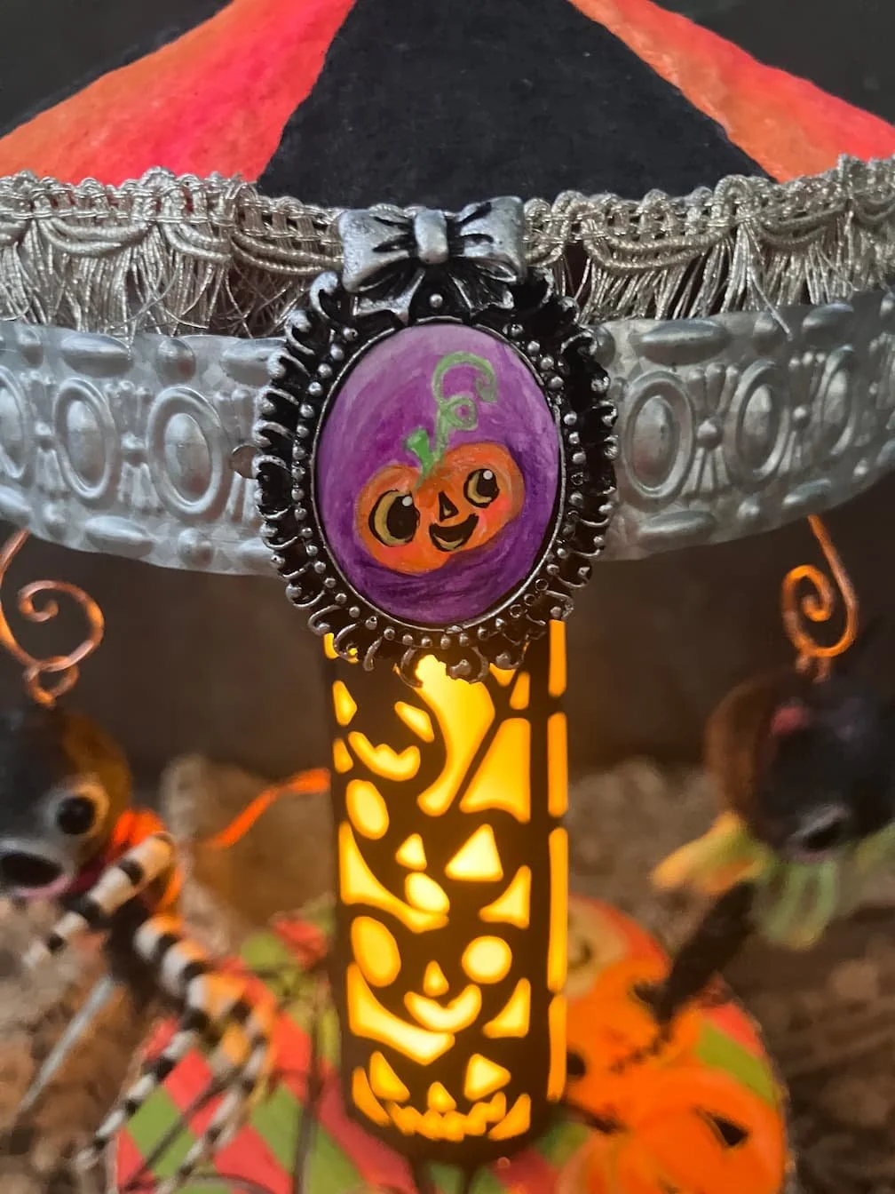 Close-up of a Halloween-themed decorated bicycle miniature with a pumpkin pendant, illuminated by a yellow light, and two small dog figurines dressed in Halloween costumes in the background.