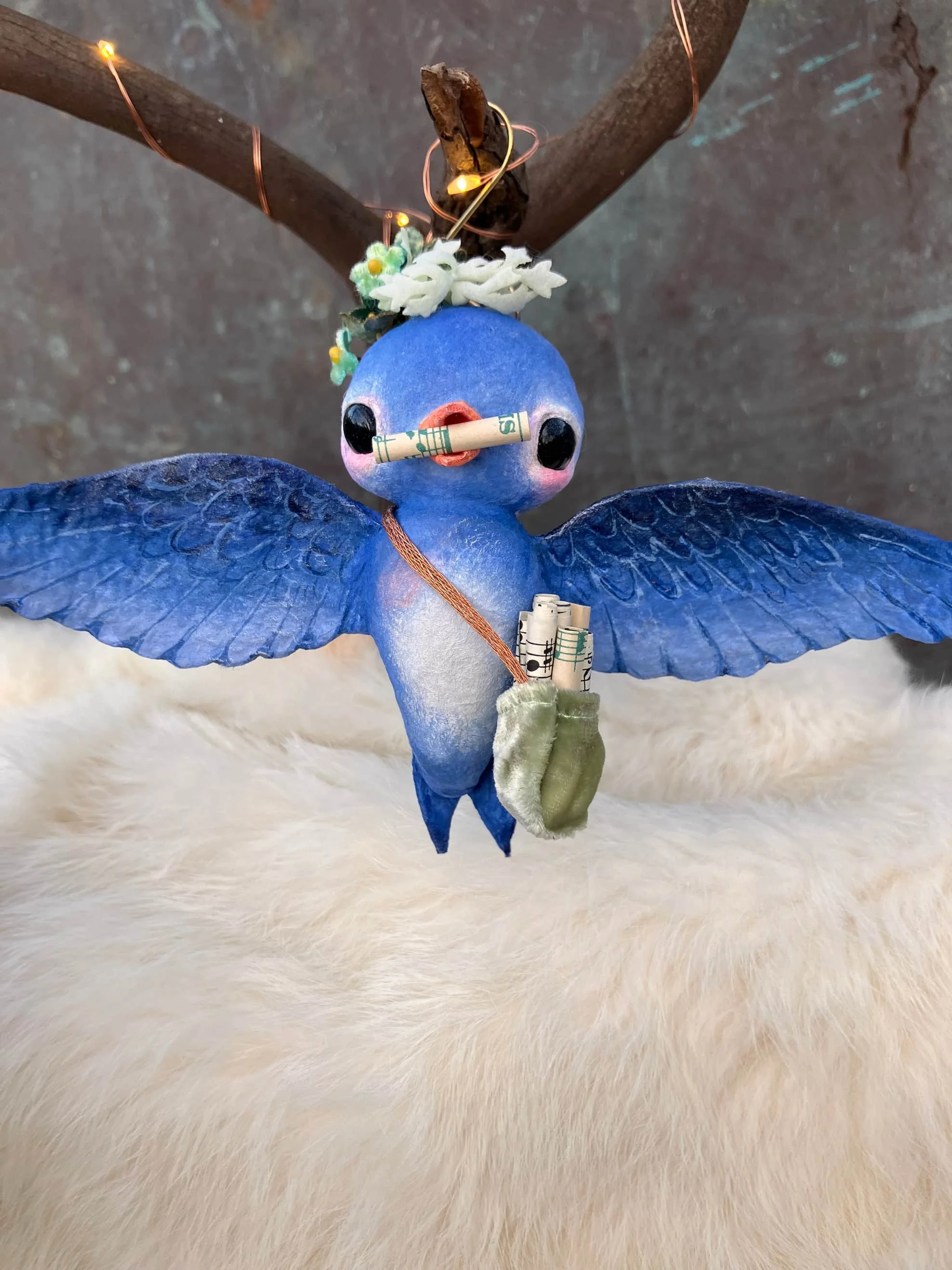 A blue bird-shaped ornament made from spun cotton with black eyes and an orange beak, holding rolled-up dollar bills in its beak and a small green sock in its wing, standing on a fluffy white surface with wooden branches and fairy lights above.