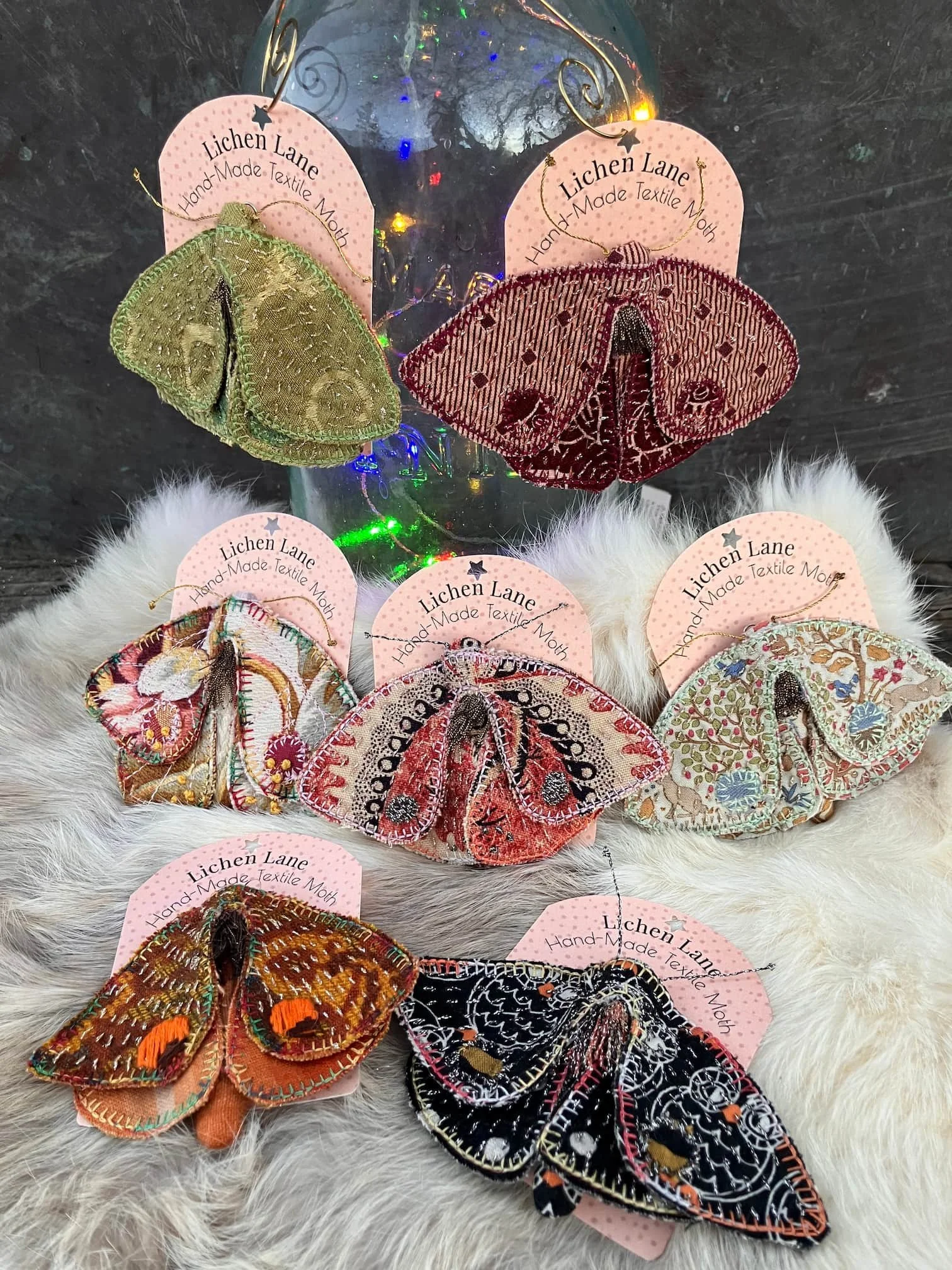 Set of embroidered butterfly ornaments hanging on pink tags labeled 'Lichen Lane Hand-Made Textile Moth,' with some ornaments featuring metallic and colorful embroidery, displayed on a white furry surface with a glass jar in the background.