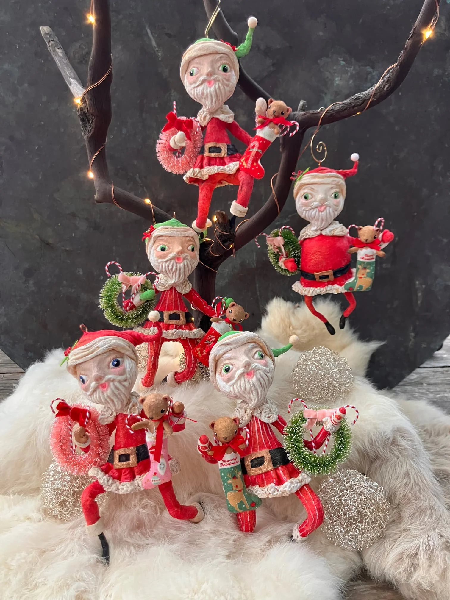 Christmas decorations featuring Elf on the Shelf characters dressed in festive outfits, teddy bears, and holiday ornaments arranged on a white furry surface with a dark background.