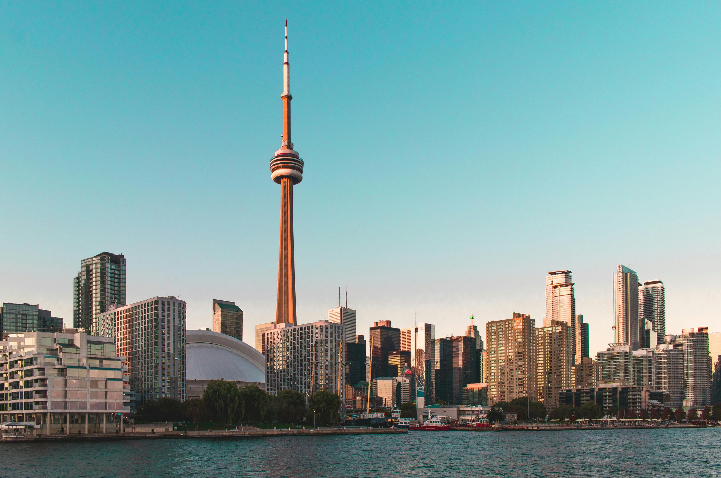 Skyline of Toronto with CN Tower in center, various skyscrapers, and waterfront in foreground during sunset.
